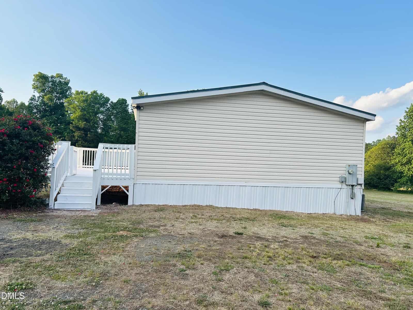A white manufactured home with a small front porch and steps, set on a dry grassy yard. Trees and bushes are in the background under a clear blue sky.