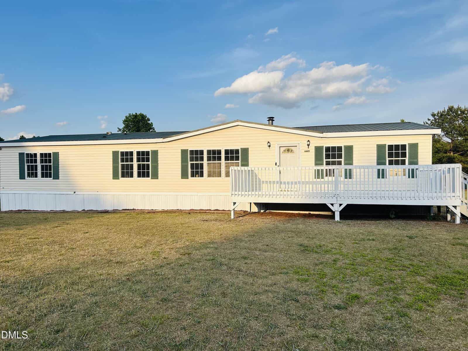 A beige manufactured home with green shutters and a white front porch sits on a grassy lawn under a blue sky with a few clouds.