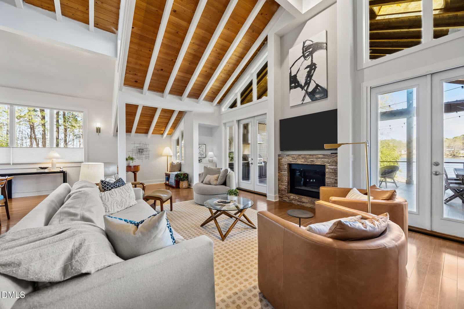 Spacious living room with vaulted wood ceiling, large windows, light gray sofa, brown leather chairs, fireplace, and glass doors leading outside. Dining area and kitchen visible in the background.