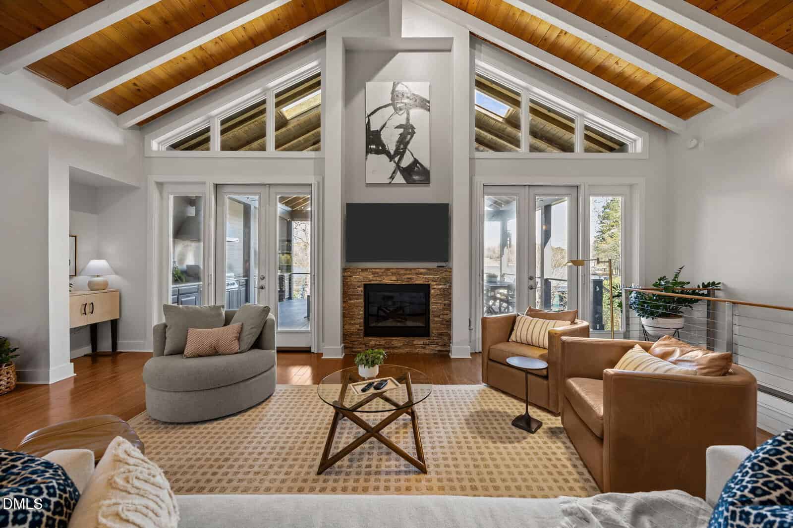 Modern living room with high wood-beamed ceiling, large windows, cozy seating, a round and a square coffee table, fireplace with stone surround, and wall art above. Natural light fills the space, creating a warm, inviting atmosphere.