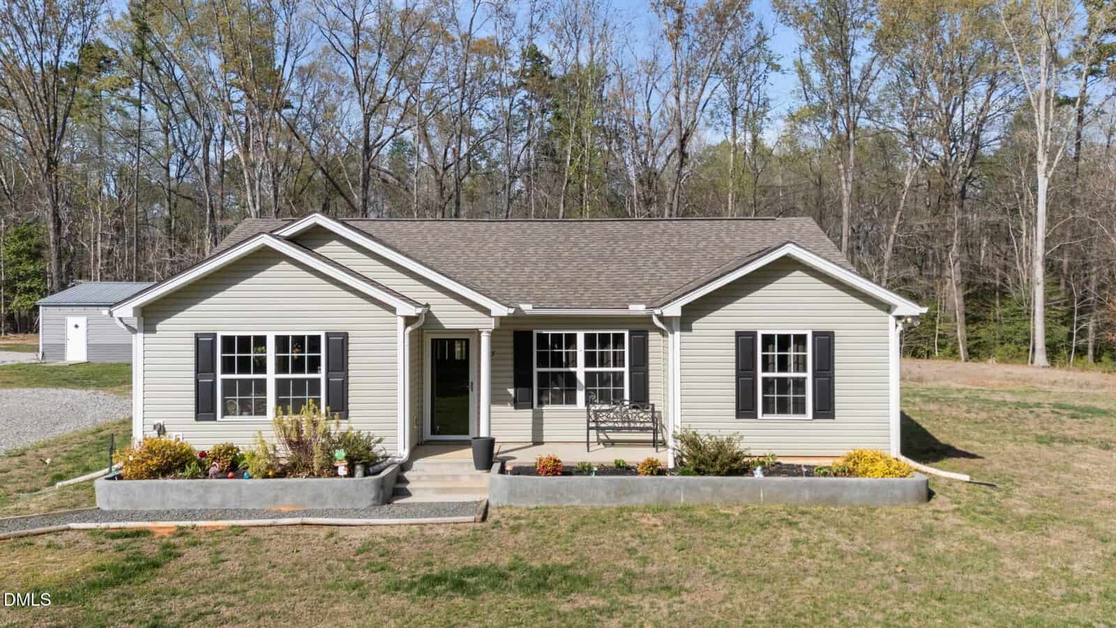 A single-story beige house with black shutters, a gabled roof, and a small front porch. The yard has low flower beds and is surrounded by grass and trees in the background.