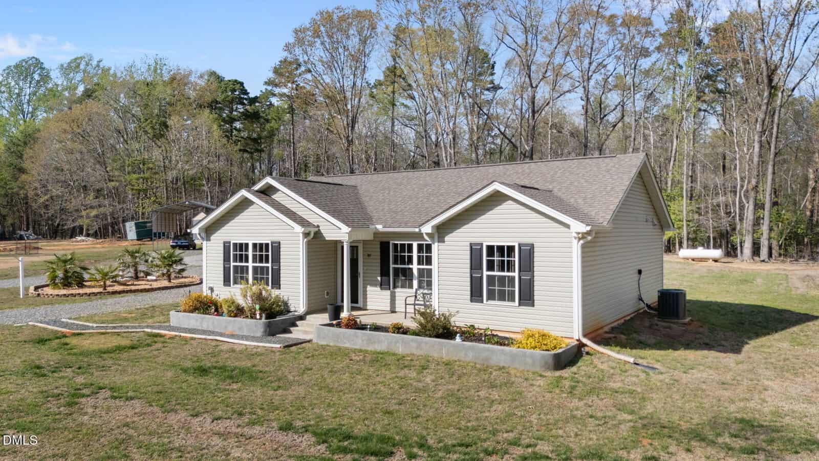 A single-story beige house with white trim and a gable roof sits on a grassy lot, bordered by trees in the background. There are rectangular flower beds in front of the house and a central walkway leading to the entrance.