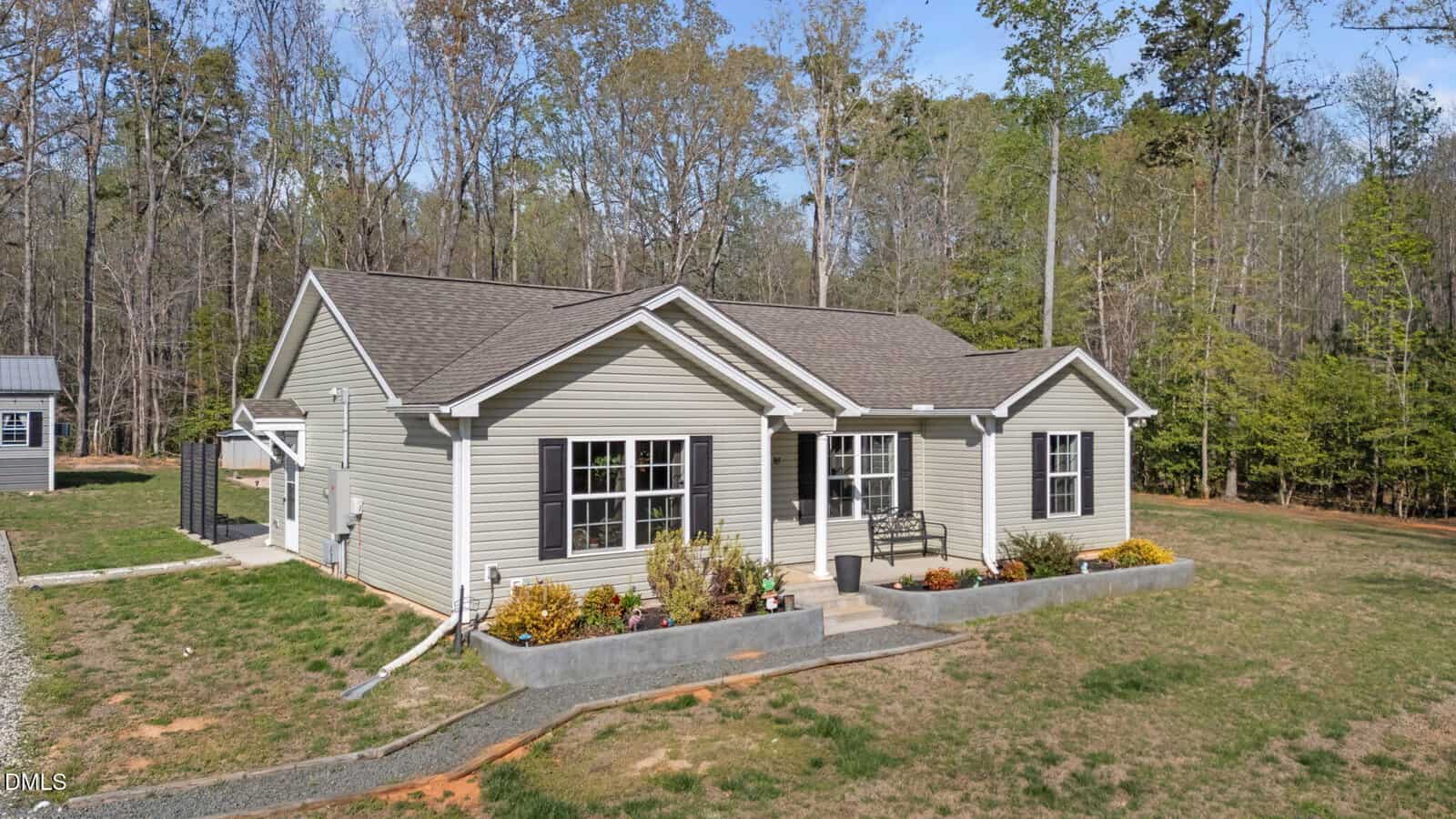 A single-story gray house with white trim sits on a grassy lot, surrounded by trees. There are several windows, a small front porch with a bench, and landscaped flower beds along the front of the house.