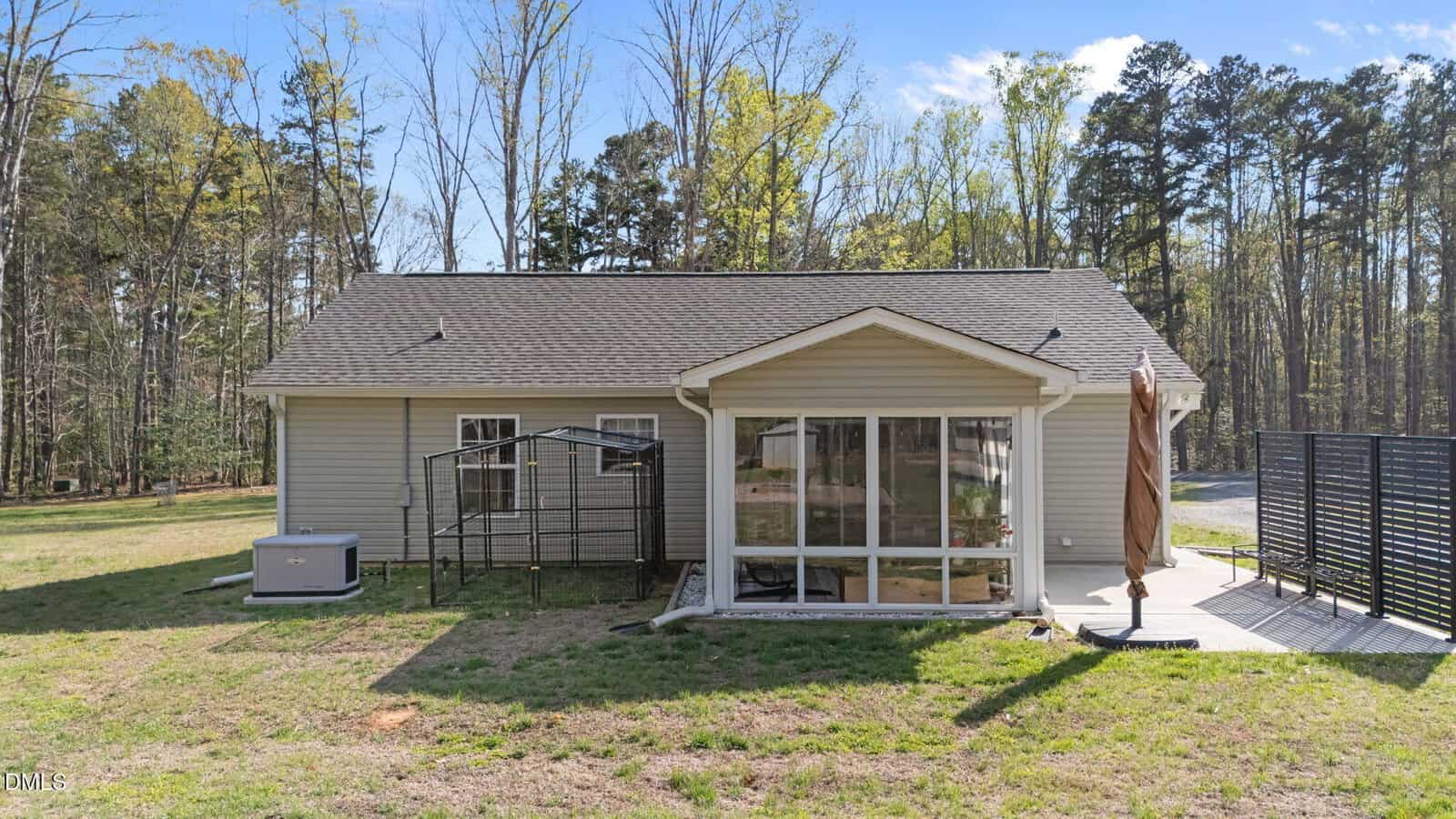 A single-story house with beige siding, a shingled roof, and a sunroom. The backyard includes a metal structure, a central air unit, an umbrella, and a black privacy screen, surrounded by grass and trees.