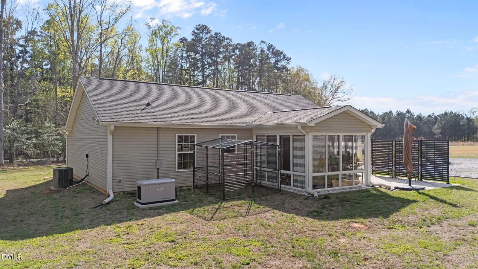A single-story beige house with a gable roof, screened porch, generator, and a metal structure in the backyard, set on a grassy lot bordered by trees under a clear blue sky.