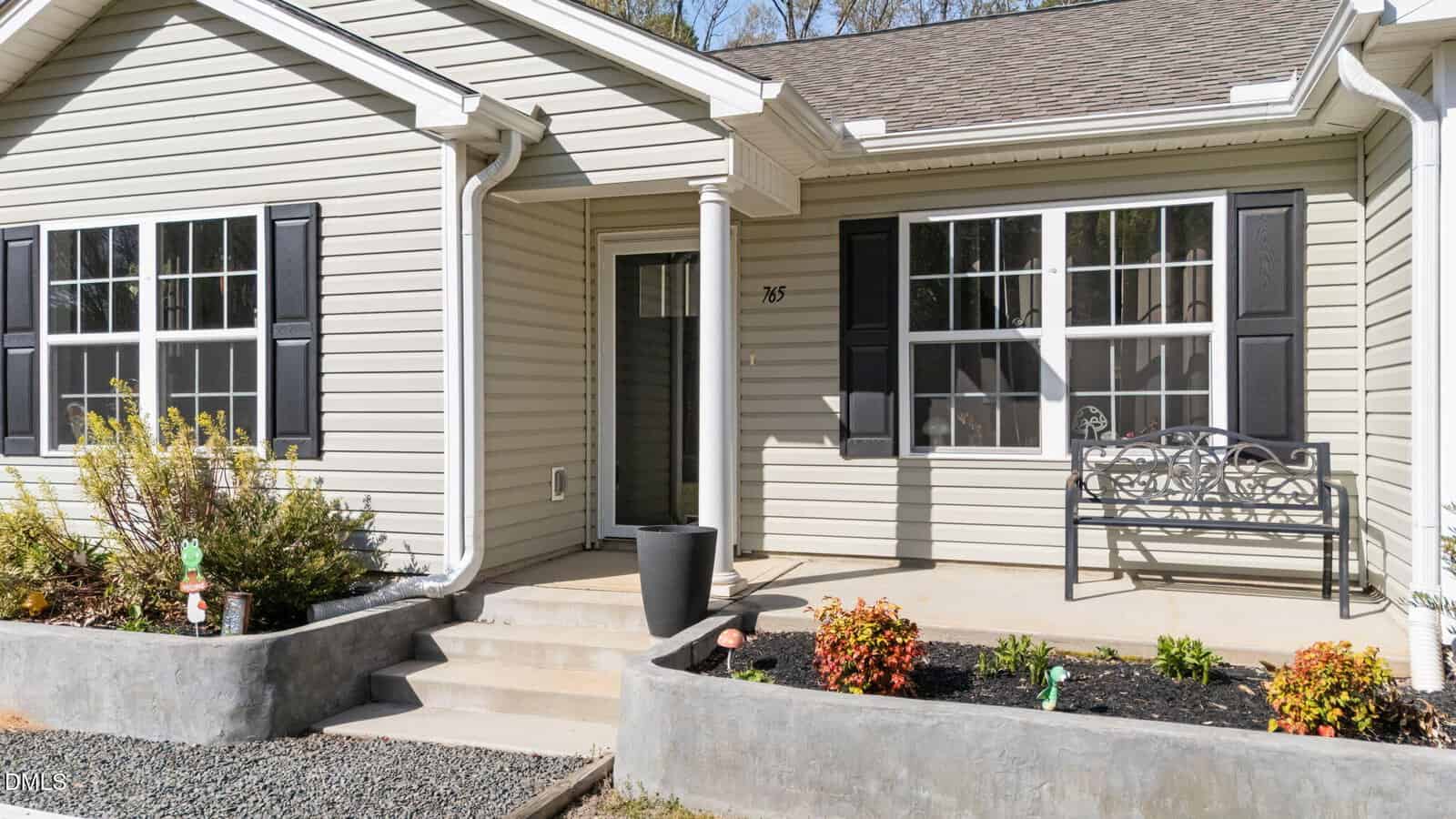 Single-story house with beige siding, black shutters, and a front porch. There are steps leading to the front door, a black planter, a metal bench, and landscaped bushes and flowers in front of the house.