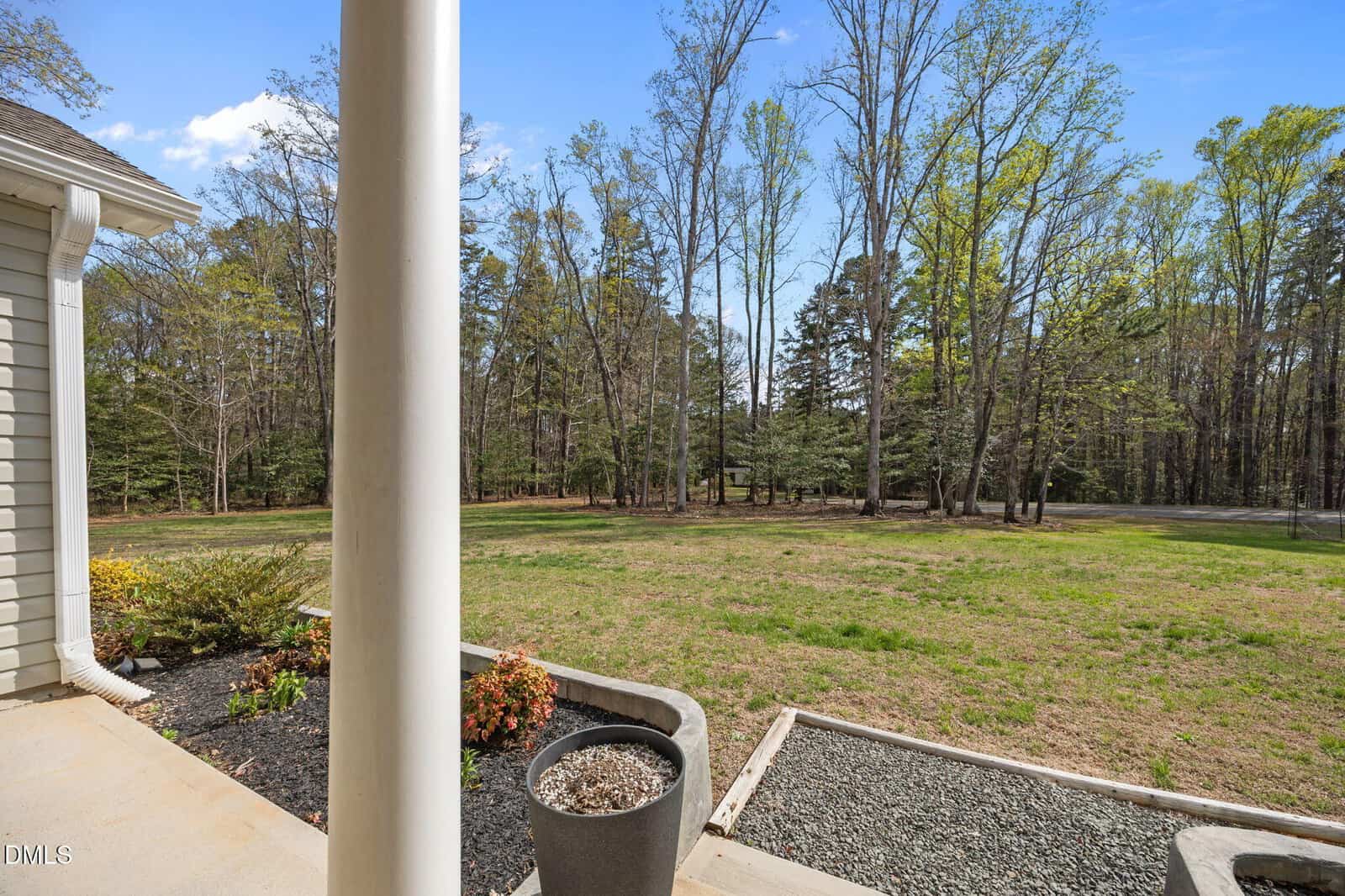View from a porch with a white column, potted plants, and landscaped area, overlooking a grassy yard bordered by tall trees under a blue sky with scattered clouds.