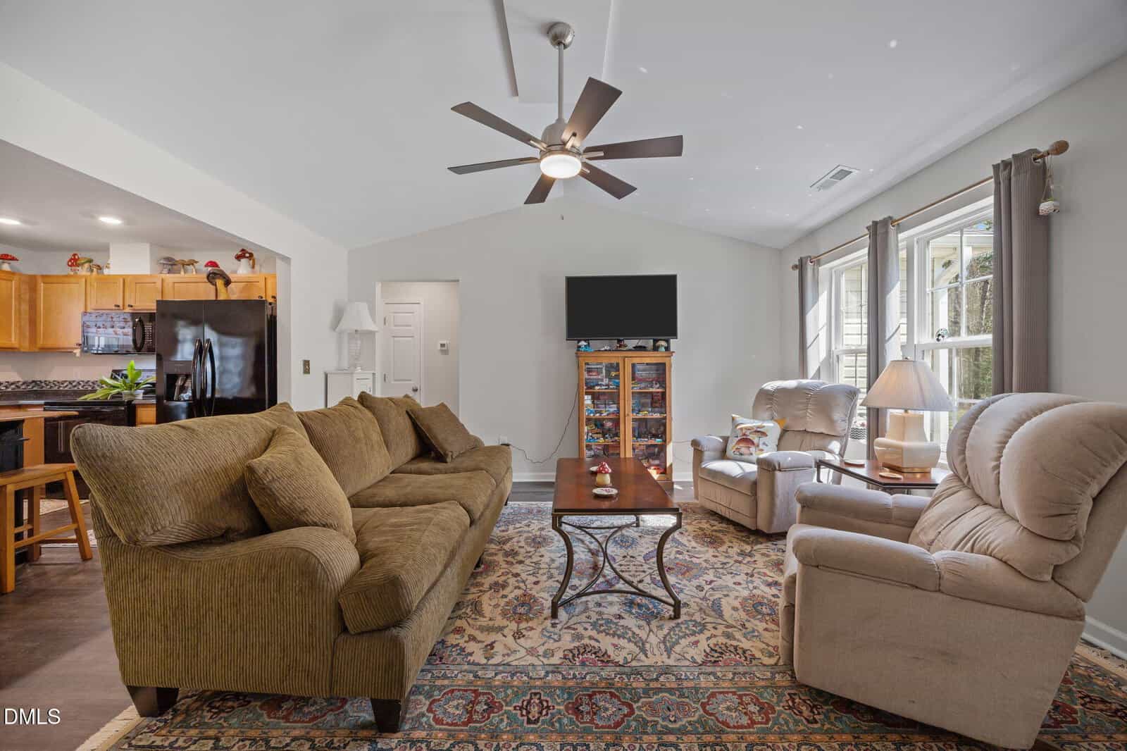 A cozy living room with a brown sofa, two beige recliners, a patterned rug, a coffee table, a TV on a stand, and large windows. The kitchen with wood cabinets is visible in the background.