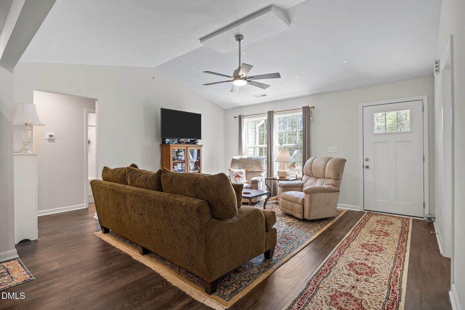 A cozy living room with brown sofas, armchairs, a TV on a stand, large windows with curtains, hardwood floors, and patterned rugs. A ceiling fan hangs overhead and light streams in through the windows and door.