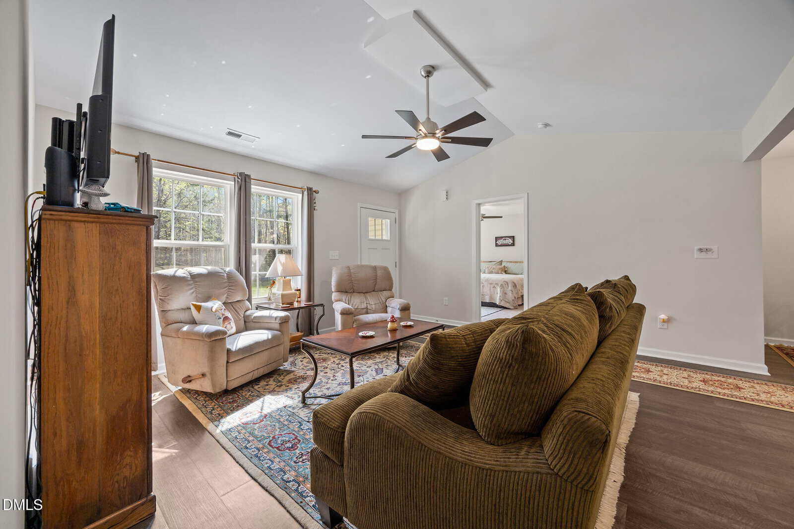 Living room with brown sofa, two beige recliners, wooden TV stand, coffee table, large window with curtains, ceiling fan, and view into a bedroom and hallway. Natural light fills the space.