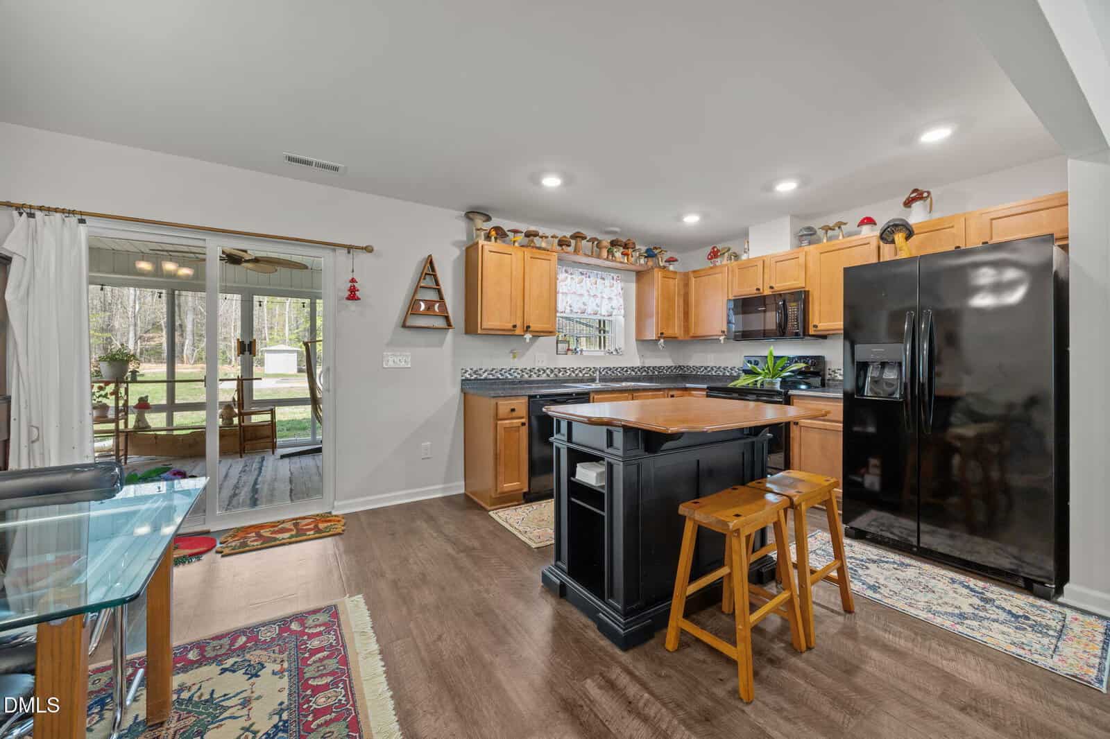 Bright kitchen with wooden cabinets, black appliances, a small island with two stools, and rugs. Large windows and a door lead to a sunroom with outdoor views. Decorative items line the upper cabinets.