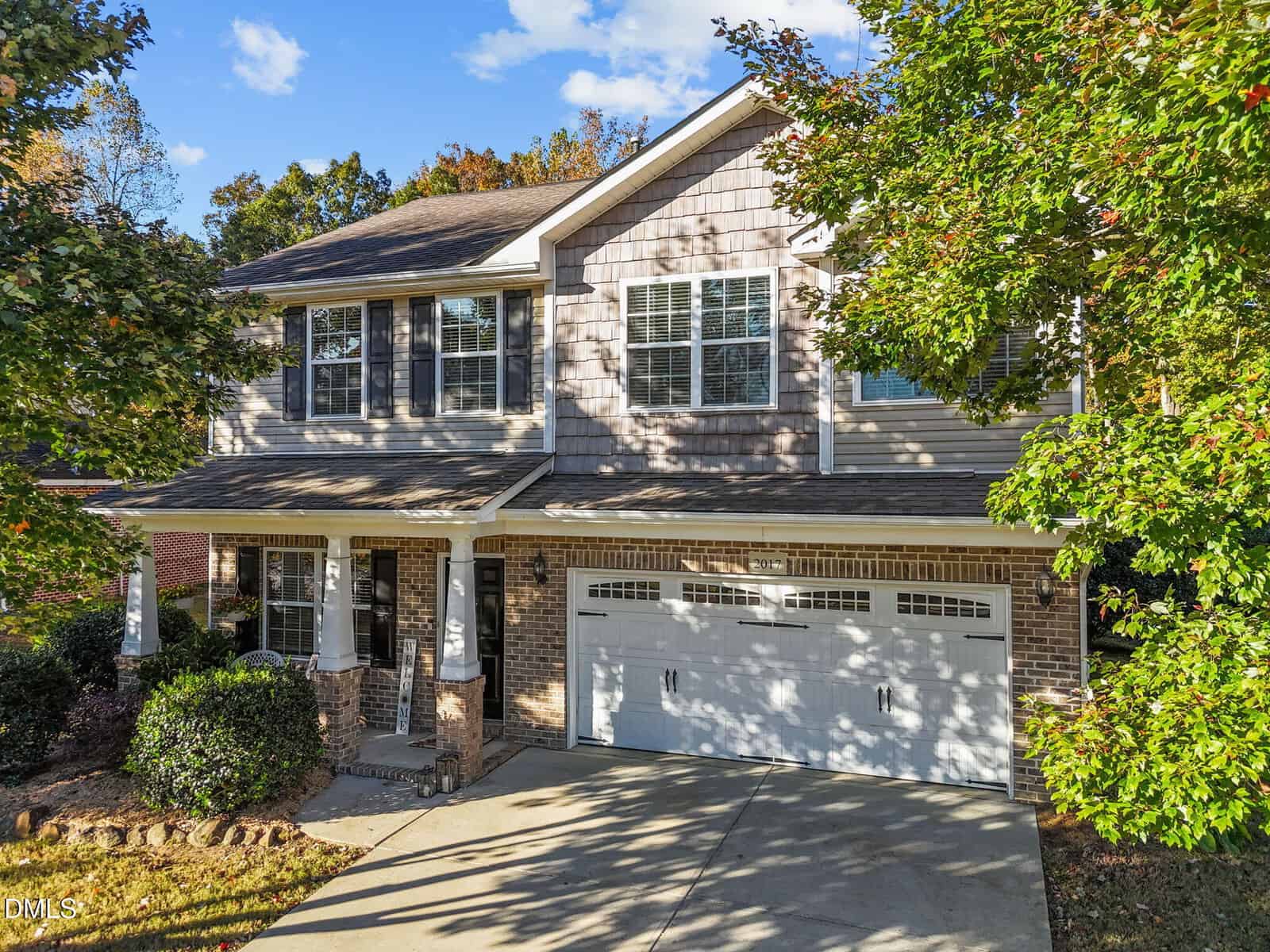 Two-story suburban house with brick and siding exterior, a covered front porch, white garage door, and driveway. Green shrubs and trees surround the front yard under a blue sky with some clouds.