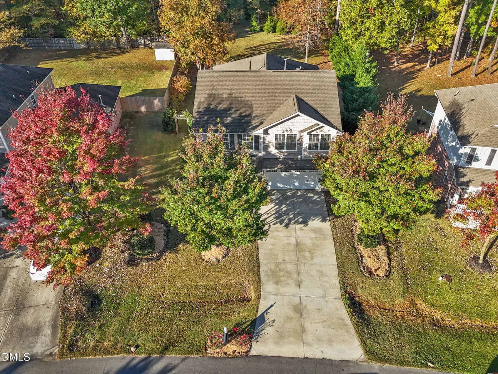 Aerial view of a single-story house with a gray roof and driveway, flanked by two trees with green and red foliage, surrounded by neighboring homes and grassy yards in a suburban neighborhood.