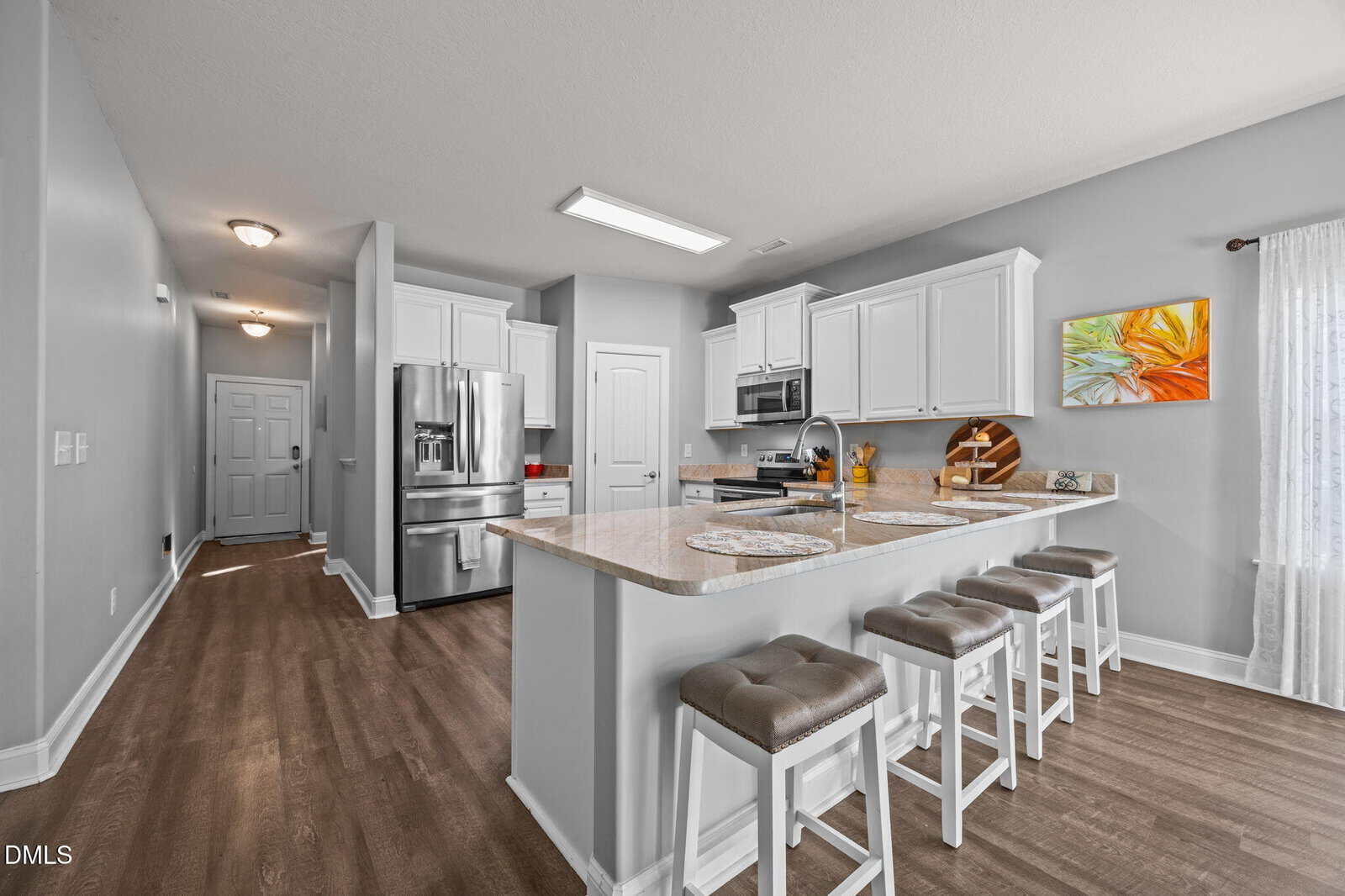 Modern kitchen with white cabinets, stainless steel appliances, light countertops, and a breakfast bar with four cushioned stools. A colorful painting hangs on the wall above the counter near a window with sheer curtains.