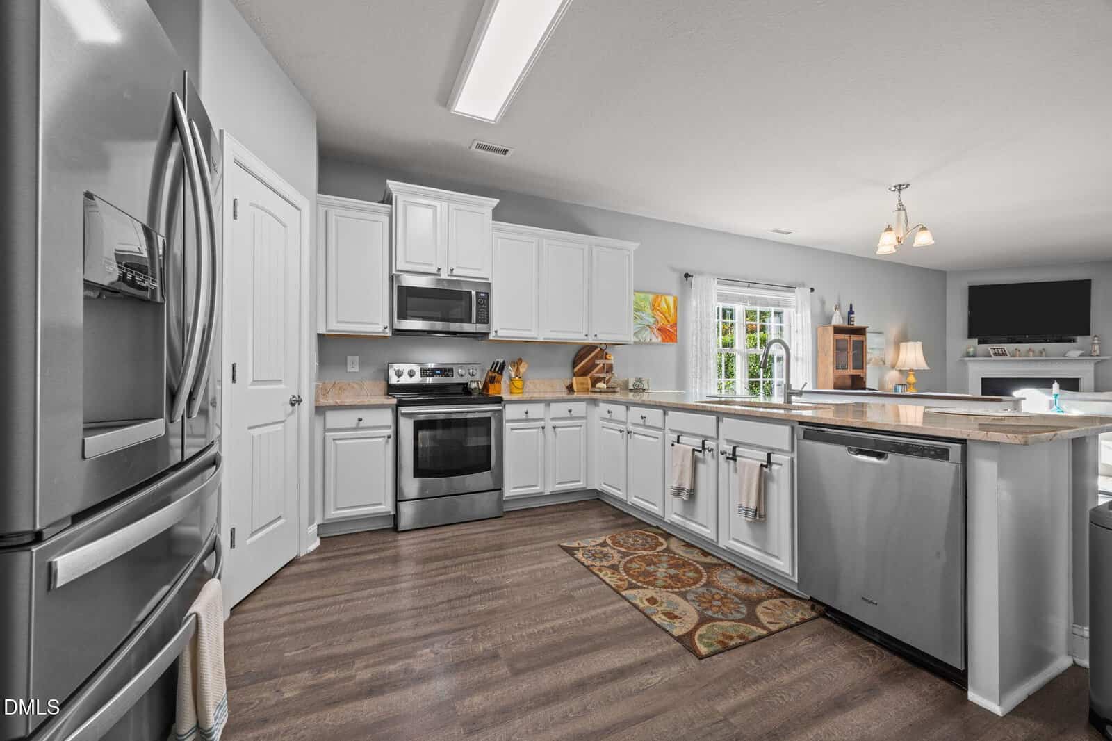 Modern kitchen with white cabinets, stainless steel appliances, a central island with sink, light wood flooring, and a patterned rug. Sliding glass doors let in natural light from the right side.