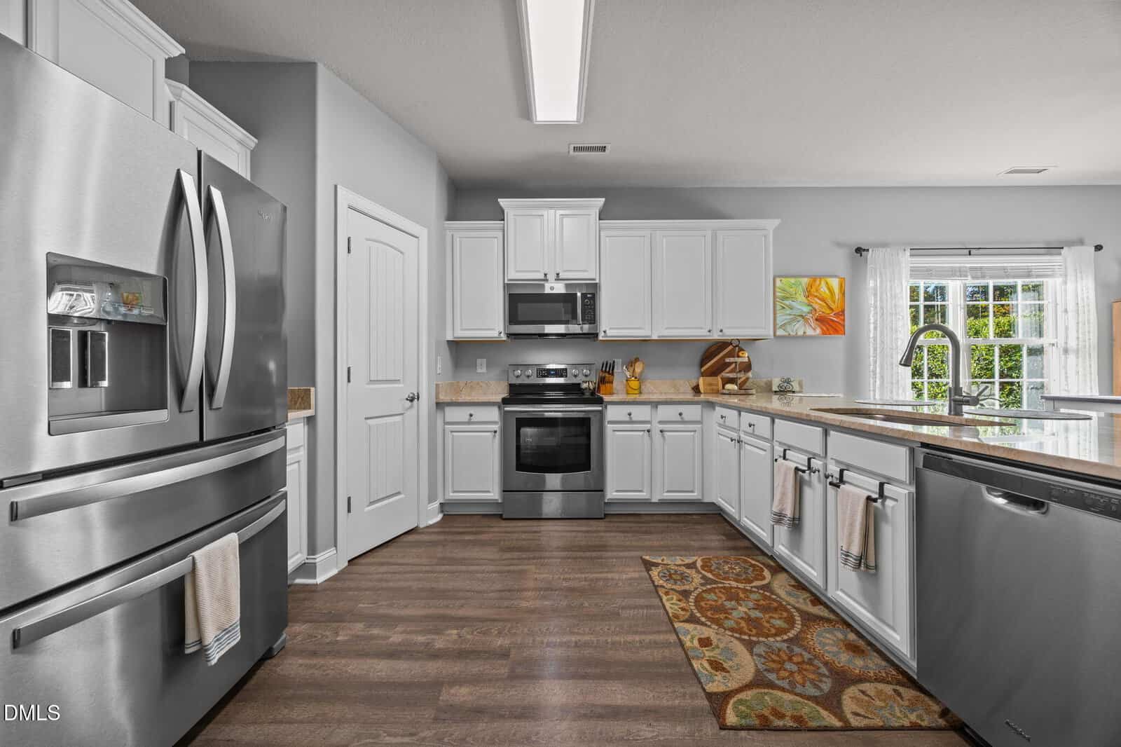 Modern kitchen with stainless steel appliances, white cabinets, a central oven, double-door fridge, dishwasher, and a window by the sink. There are decorative rugs on the wooden floor and a painting on the wall.