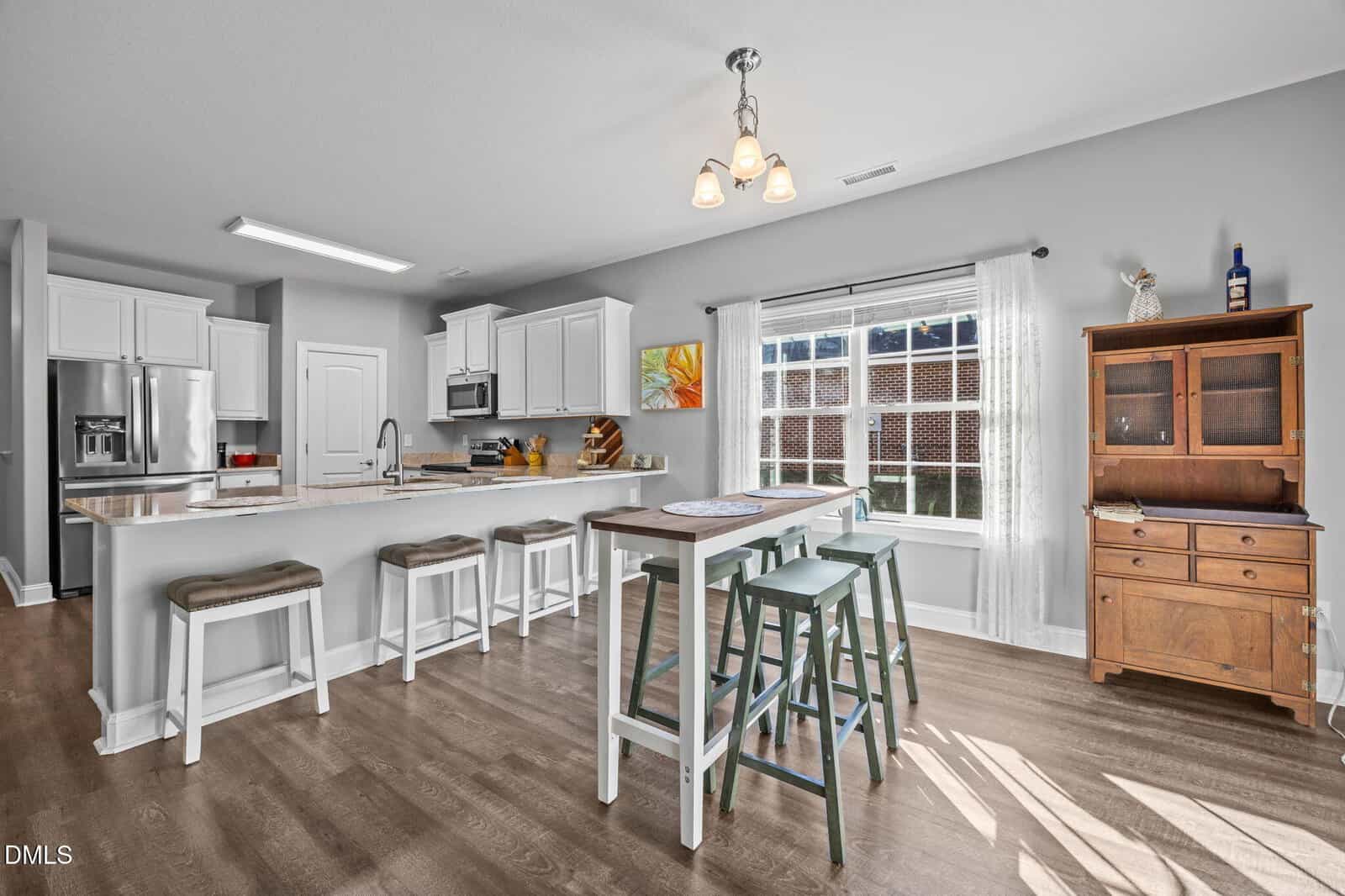Bright, modern kitchen with white cabinets, stainless steel appliances, a large island with four stools, a wooden dining table with benches, and a window letting in natural light. A wooden hutch stands against a gray wall.