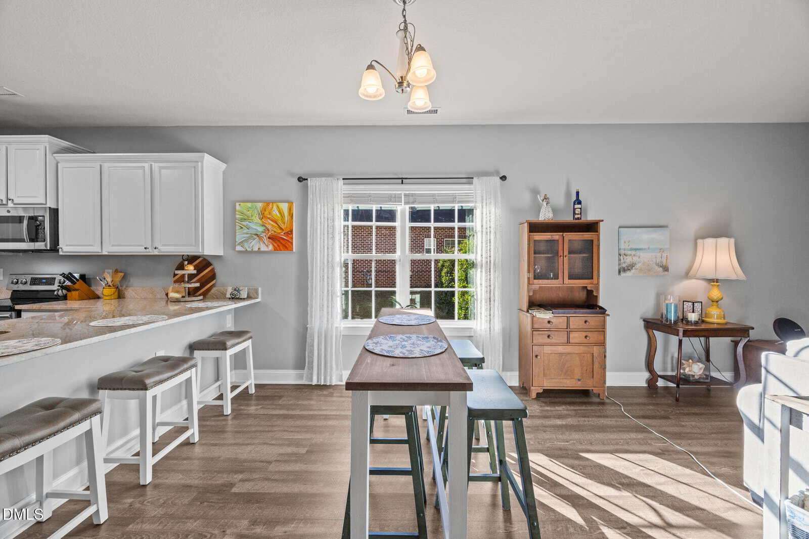 Bright kitchen and dining area with white cabinets, a wooden table with stools, a large window with sheer curtains, wall art, and a wooden hutch in a cozy, modern home. Light wooden flooring throughout.