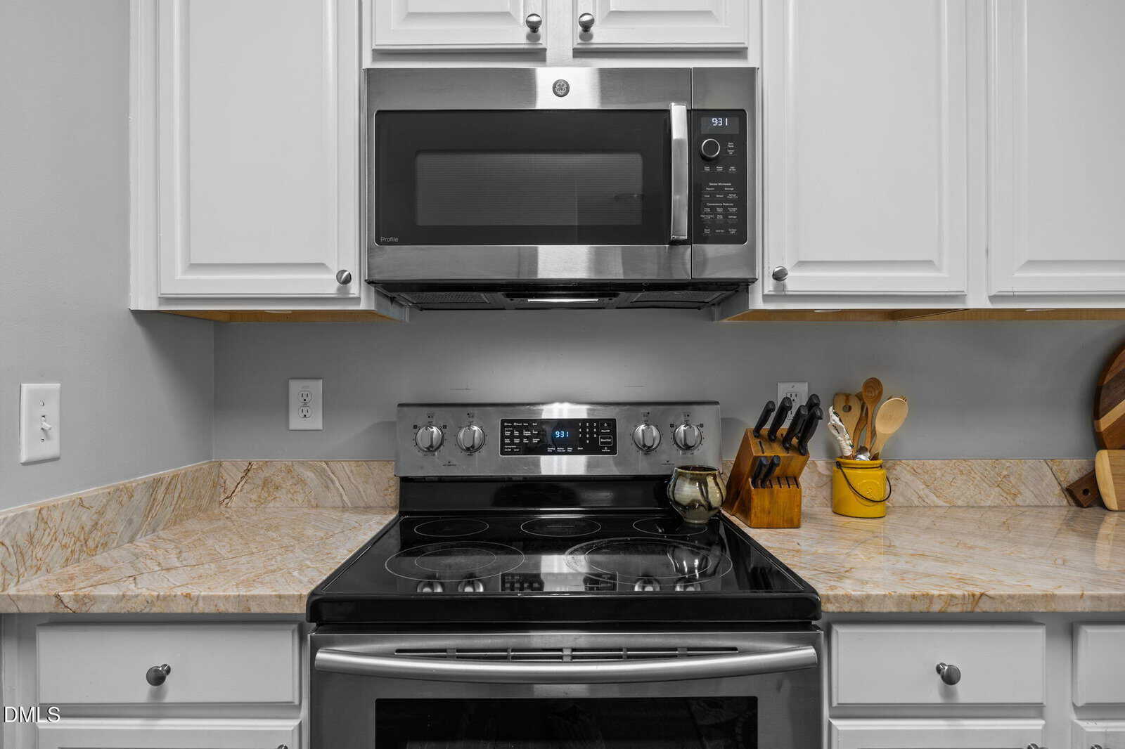Modern kitchen with a stainless steel stove and microwave, white cabinets, marble countertop, a knife block, utensils in a holder, and a cutting board on the right side.