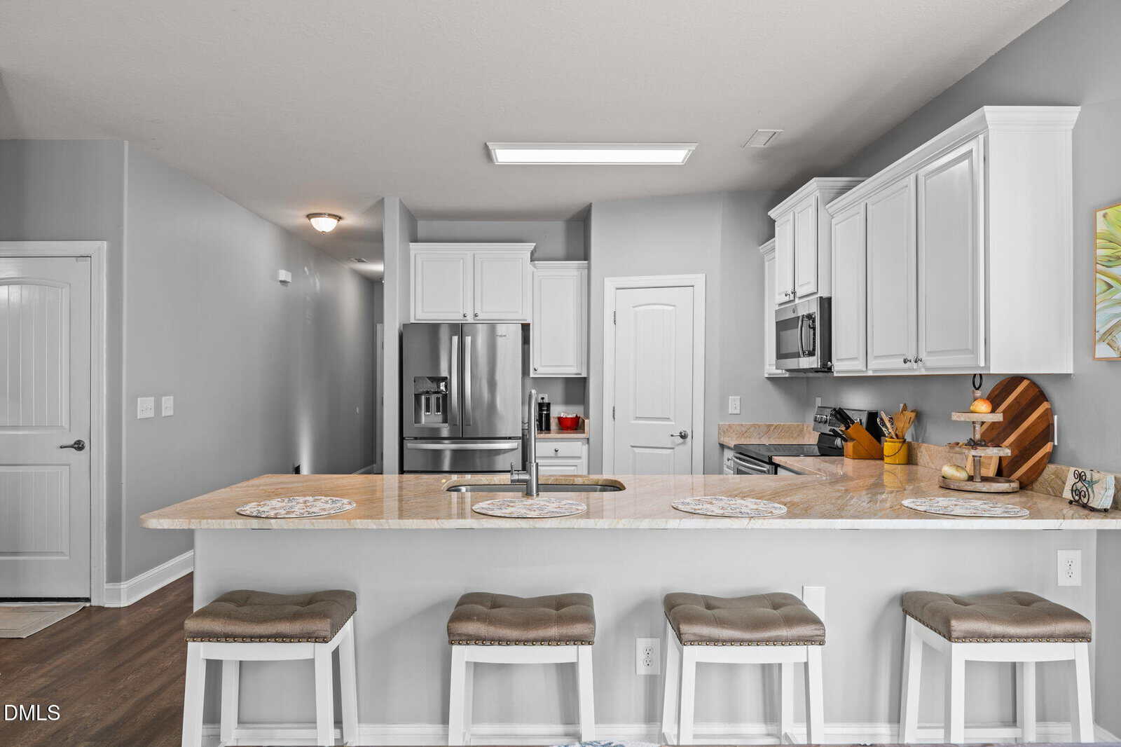 Modern kitchen with white cabinets, stainless steel appliances, beige countertops, and four cushioned stools at a breakfast bar. Walls are light gray; a cutting board and utensils are on the counter near the stove.