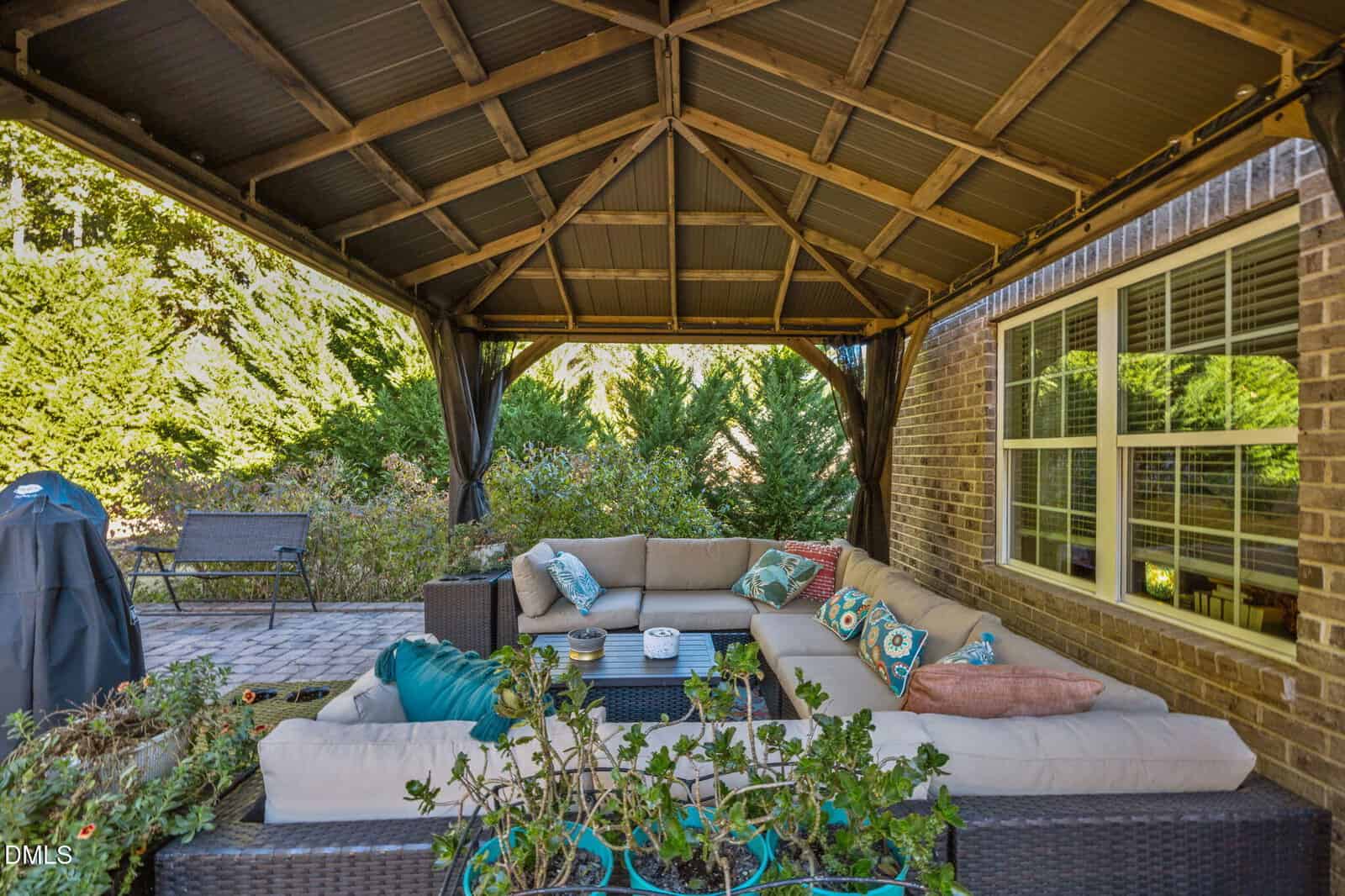A cozy outdoor patio with a large beige sectional sofa under a wooden gazebo, accented with colorful pillows. Greenery surrounds the space, and a brick wall with windows is visible on the right.