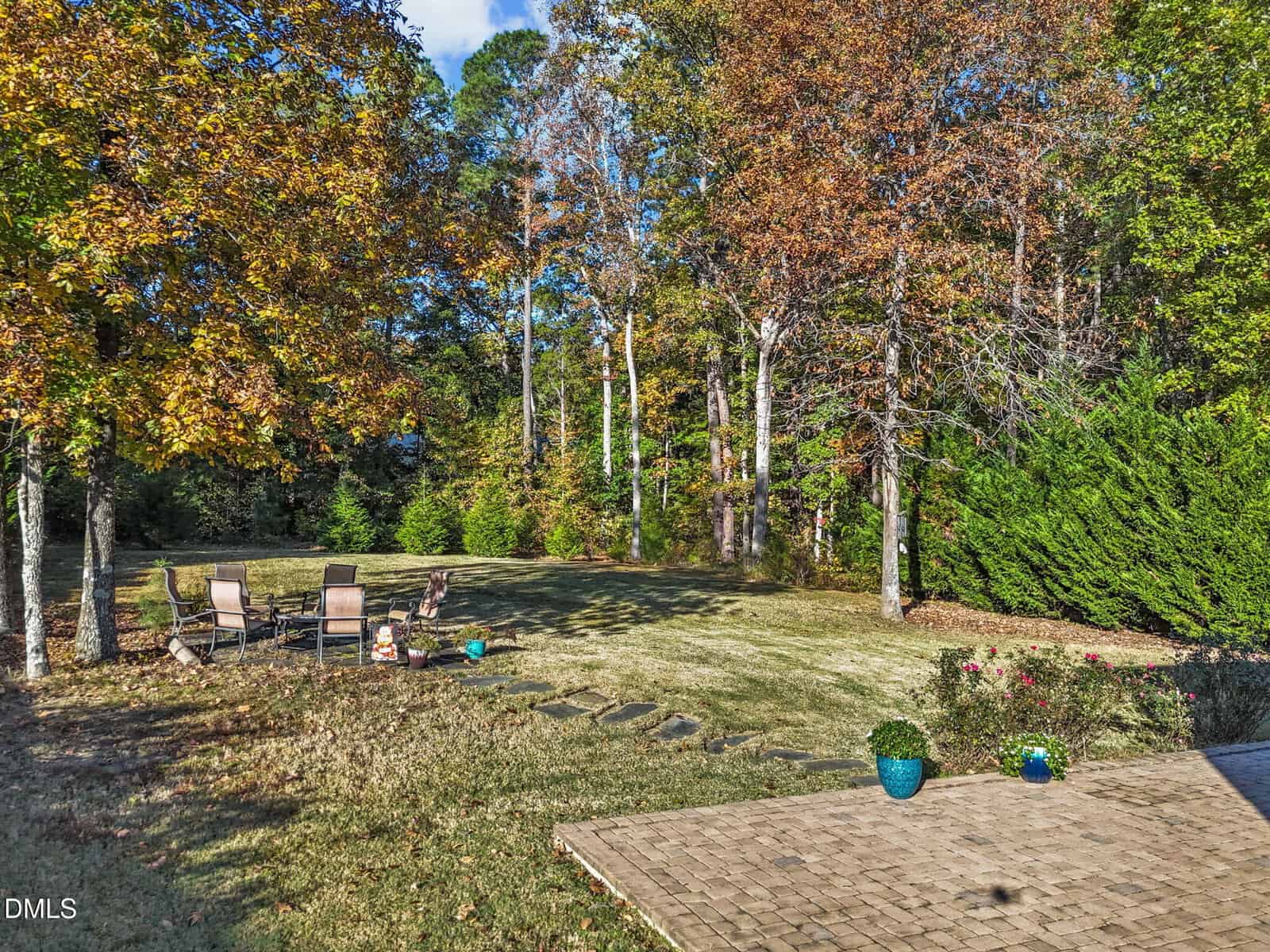 A spacious backyard with a stone patio, potted plants, a grassy lawn, and a seating area with chairs around a fire pit. Tall trees with autumn leaves border the yard under a blue sky.