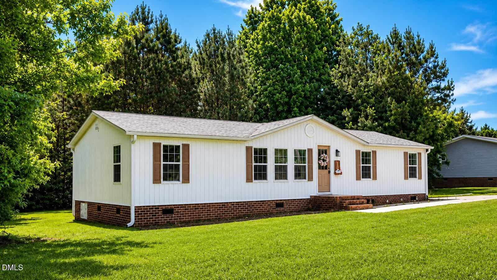 A single-story white house with brown shutters and a brick foundation sits on a well-manicured lawn at 7537 Sam Young Road, Oxford, surrounded by green trees under a bright blue sky.