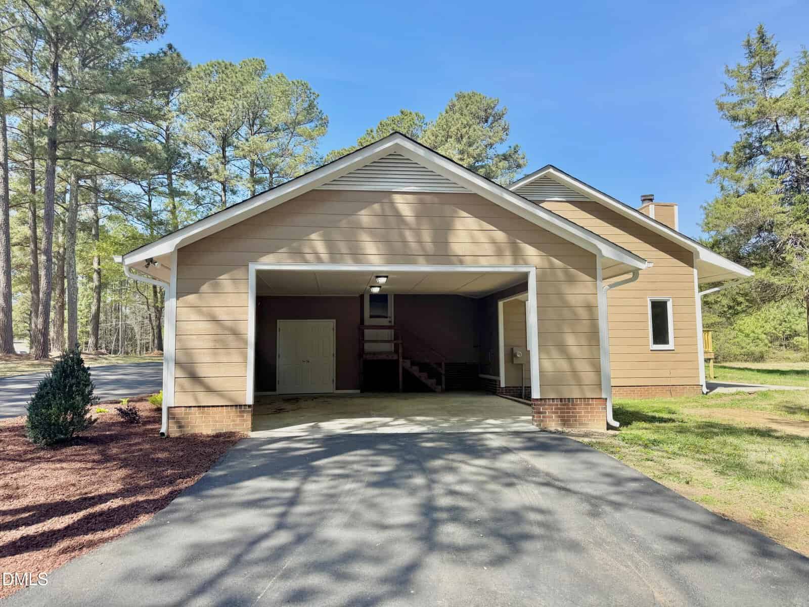 A tan house with an attached two-car garage sits among tall trees. The garage door is open, showing a staircase, a door, and an unfinished interior. The driveway leads up to the garage, and sunlight filters through the trees.