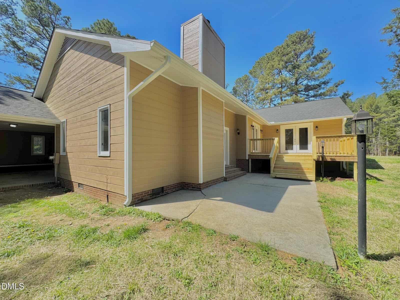 Single-story tan house with white trim features a chimney, elevated deck with railing, concrete patio, and lawn. Surrounded by trees under a clear blue sky.