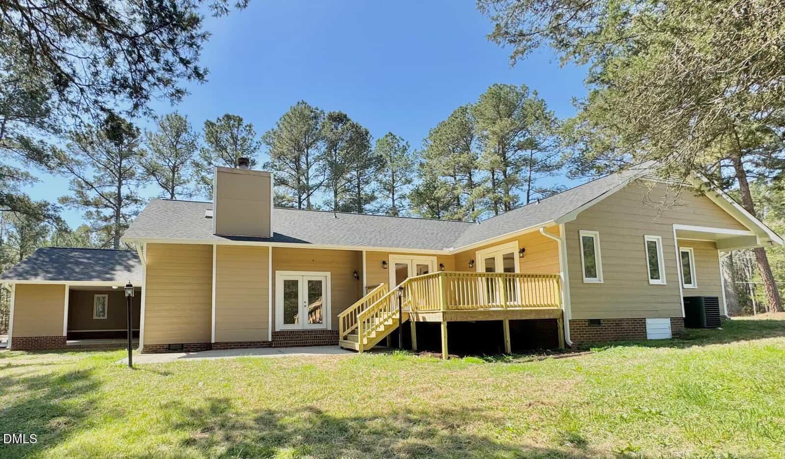 A beige single-story house with a dark roof, elevated wooden deck, and multiple windows and doors. The backyard is grassy with surrounding tall pine trees under a clear blue sky.