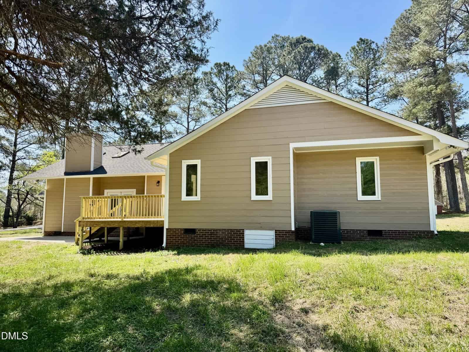Single-story tan house with white trim, three rectangular windows, a small back deck with a wooden railing, and an outdoor HVAC unit. Pine trees surround the house and the yard has green grass.