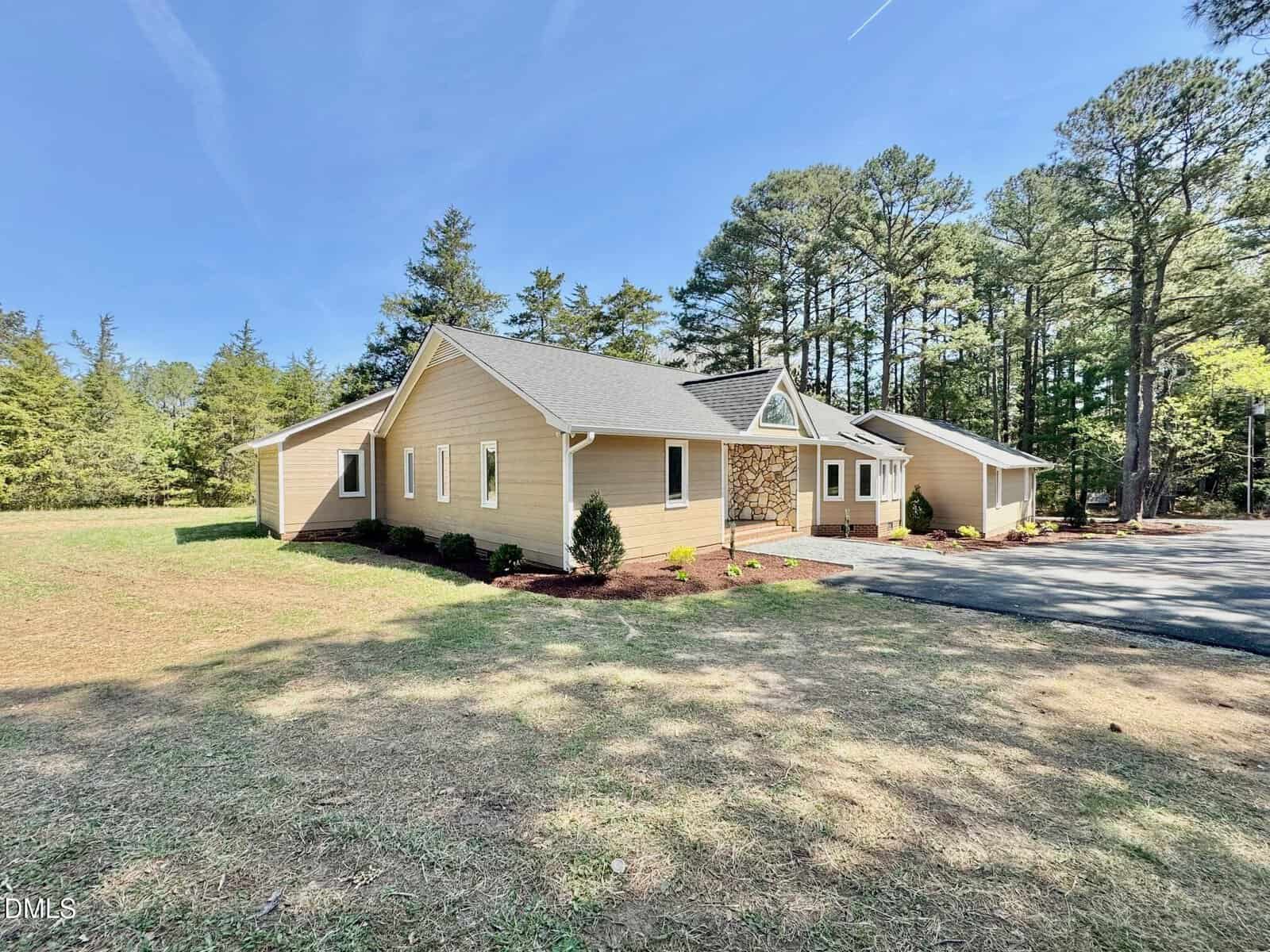 A tan, single-story house with white trim sits on a large grassy lot surrounded by tall trees under a blue sky. A paved driveway leads to the front entrance.