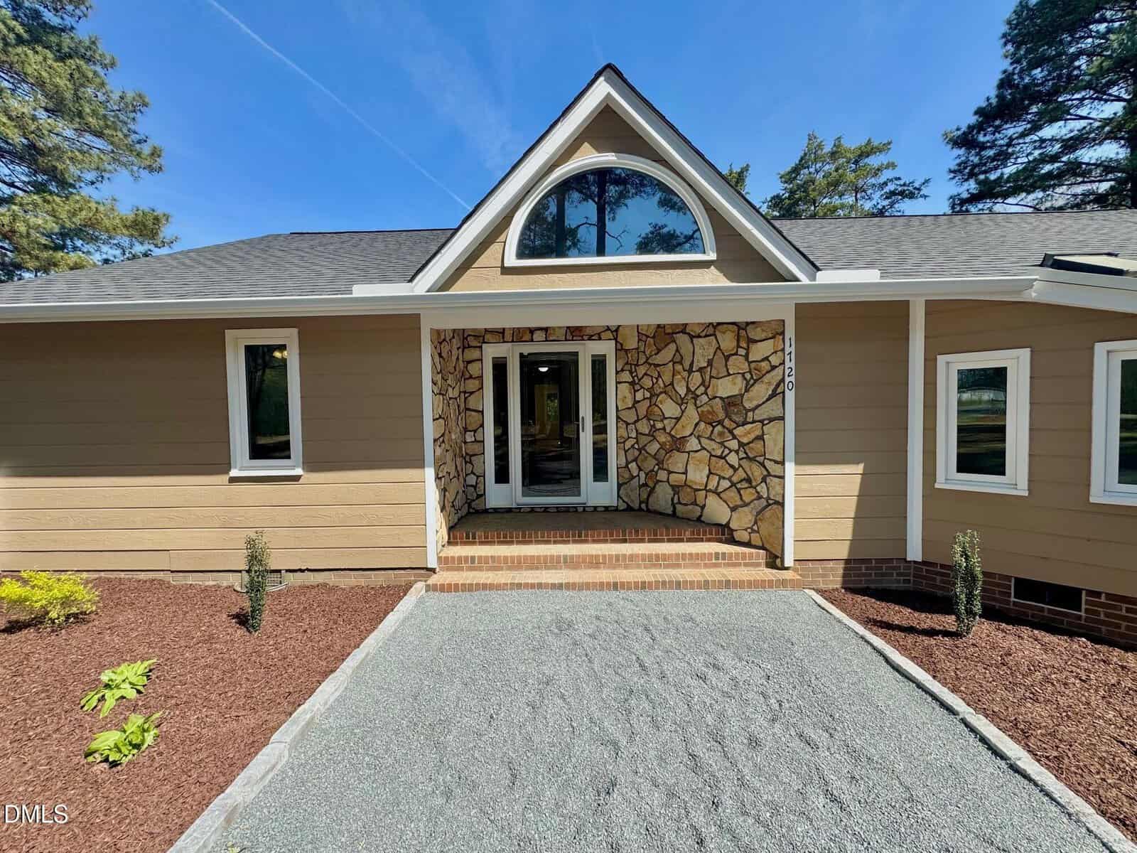 A tan house with stone accents around the entrance, large arched window above the door, gravel path, and minimal landscaping with small bushes and mulch.