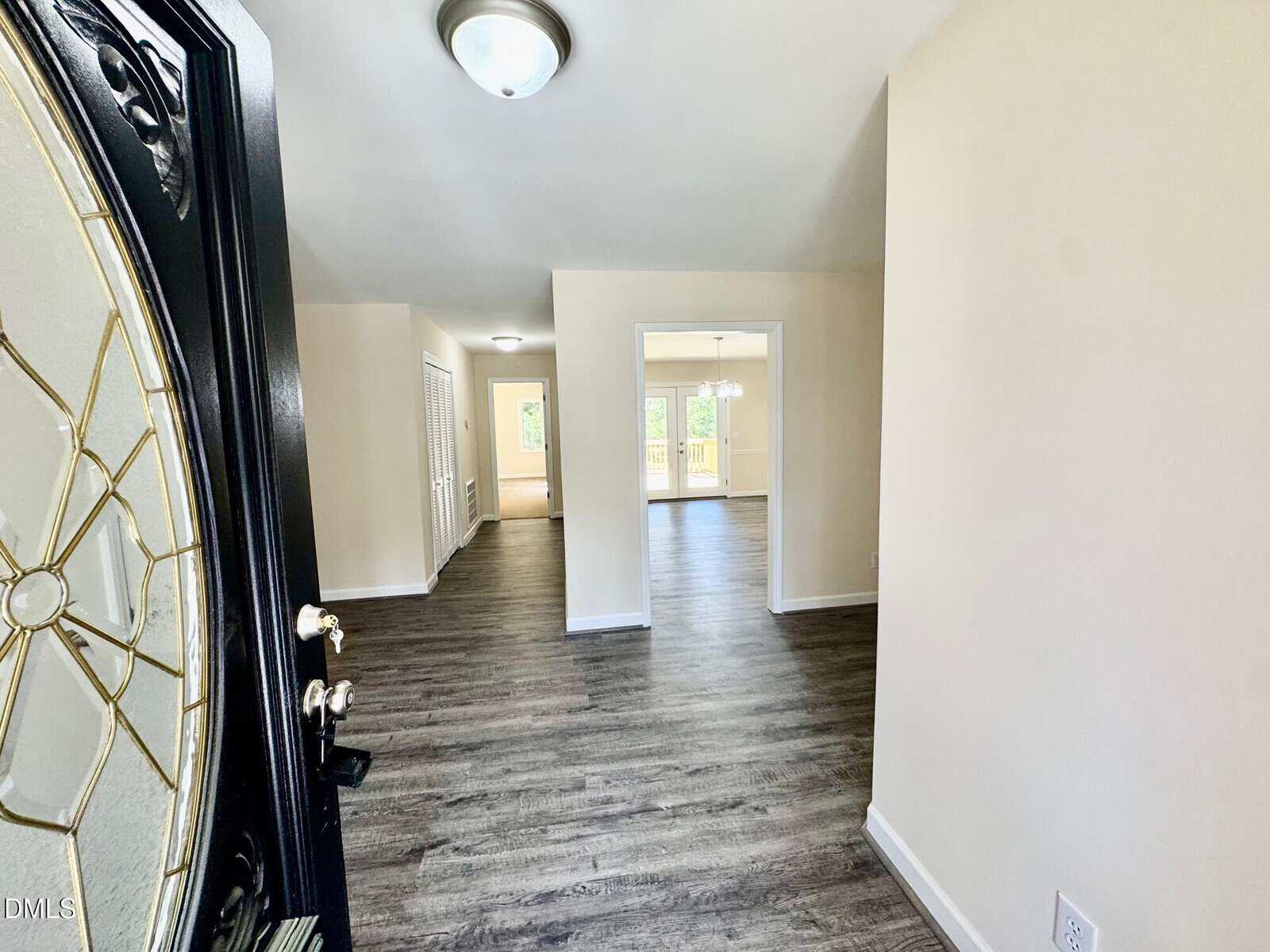 View from an open front door into a bright home with wood floors, beige walls, and an open layout leading to a back patio with glass doors. Natural light fills the space.