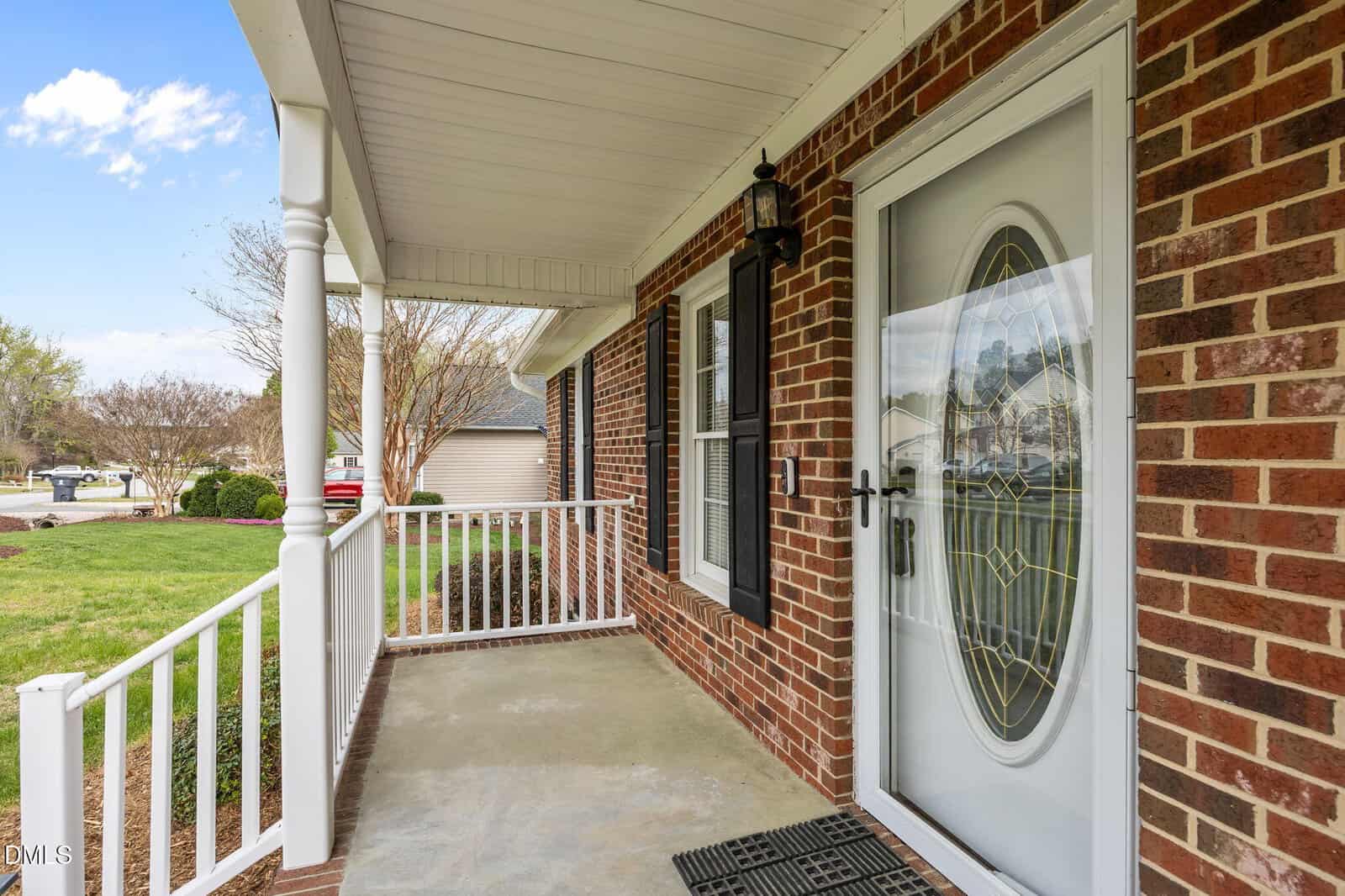 A brick house with a covered front porch, white railing, a decorative oval glass front door, and a concrete floor. A welcome mat sits near the door, and the yard with grass and driveway is visible.