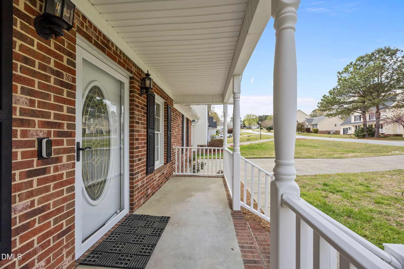A brick house with a covered front porch, white railing, two columns, a glass front door with a black lantern-style light fixture, and a view of a residential neighborhood street and yards.