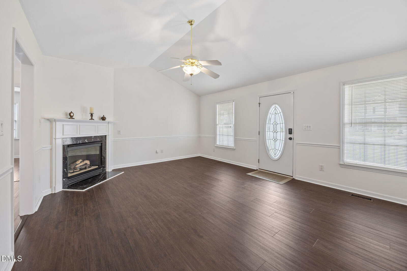 Bright living room with wood floors, white walls, a ceiling fan, fireplace with mantle on the left, oval glass front door, and large windows letting in natural light.