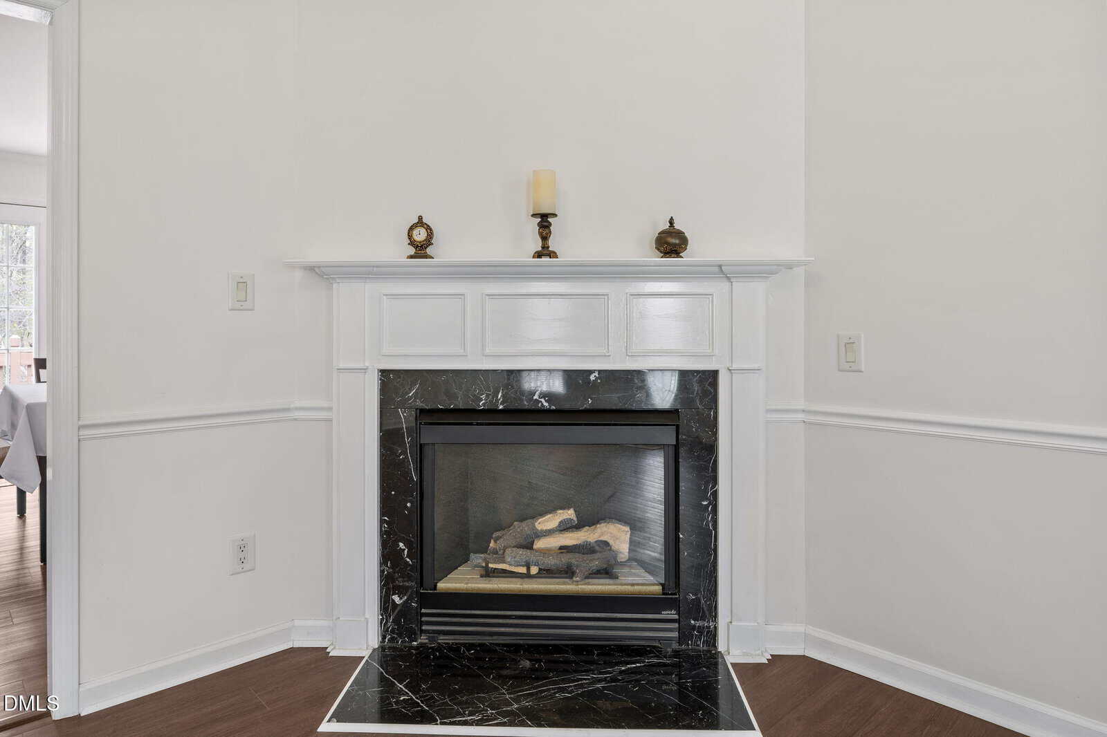 A white fireplace with a black marble surround, decorated with a candle and small ornaments on the mantel, is set against a light-colored wall with wainscoting and wood flooring.