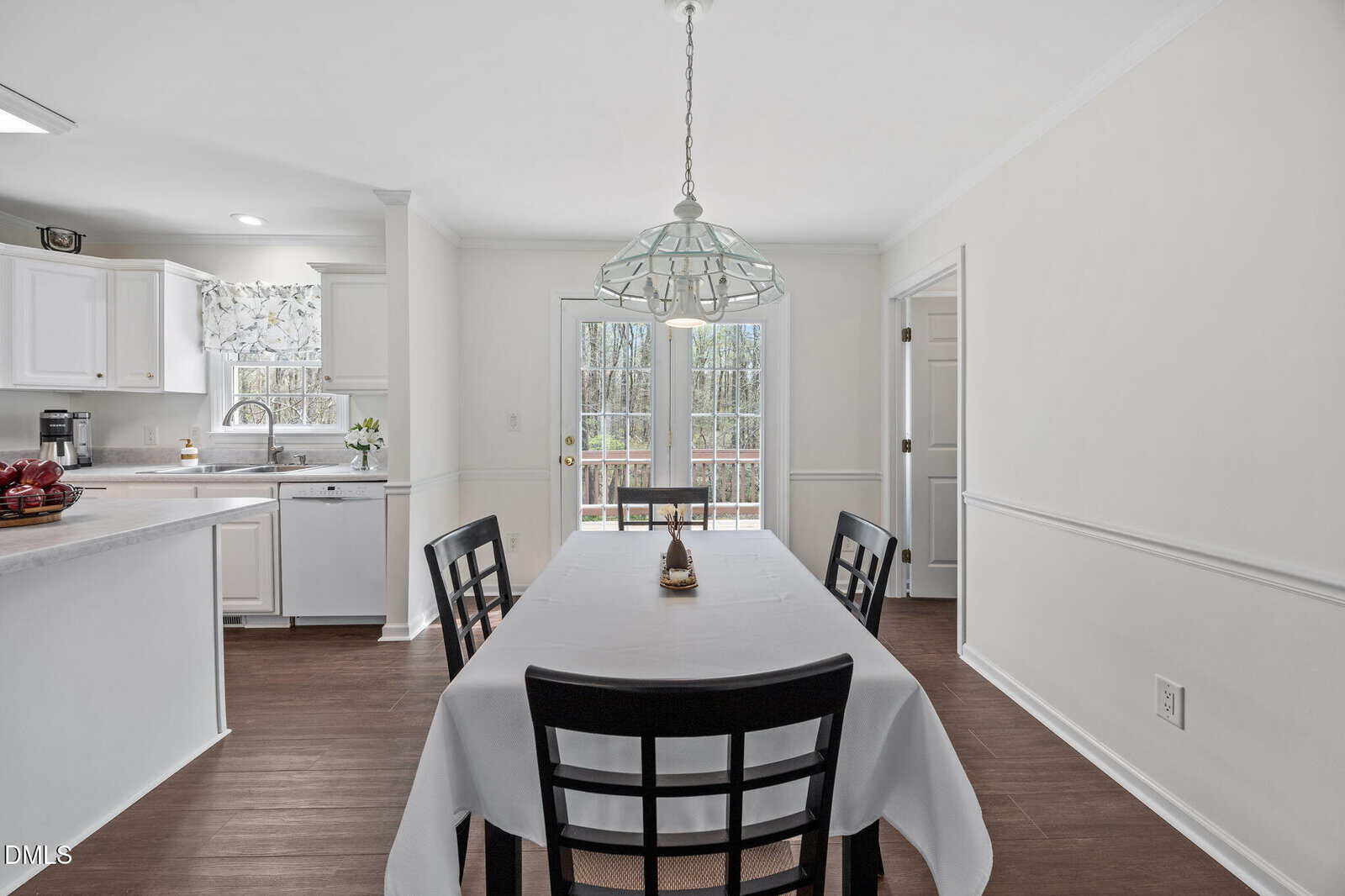 A dining area with a table for four, white tablecloth, black chairs, and a pendant light. The space opens to a kitchen on the left and glass doors leading outside at the back. Walls and cabinets are white.