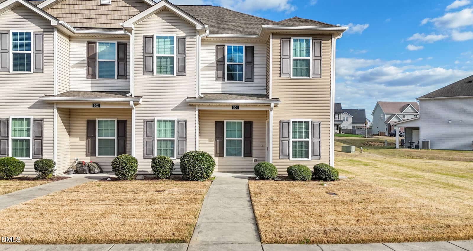 Two-story beige townhouse with white trim and brown shutters, four front windows, a covered entryway, trimmed bushes along the facade, and a sidewalk leading to the front door. Neighboring houses and a grassy yard are visible.
