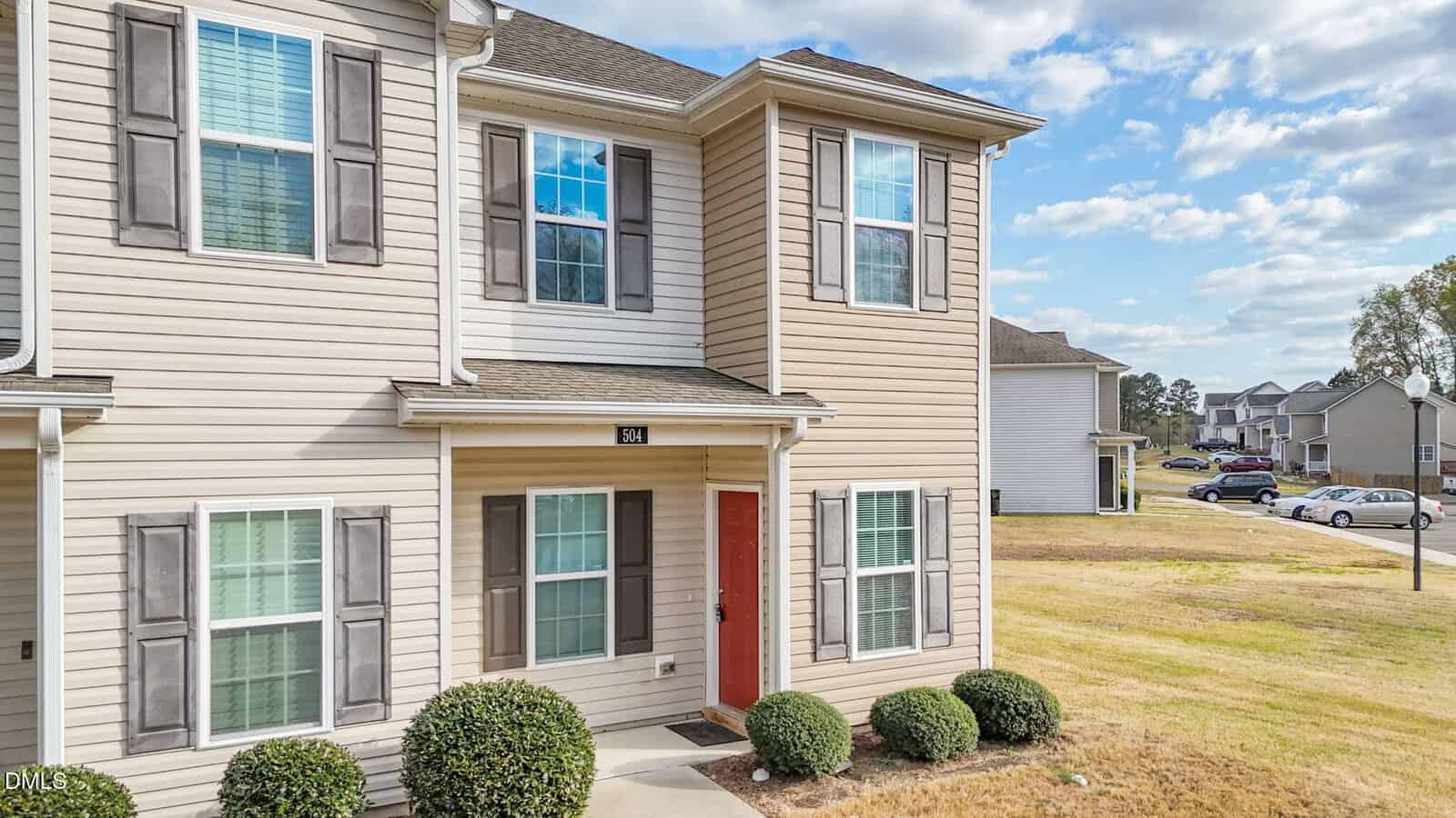 Two-story beige townhouse with gray shutters and a red front door. There are three bushes in front of the entrance, and a sidewalk leads to the door. Other similar homes and a street are visible in the background.