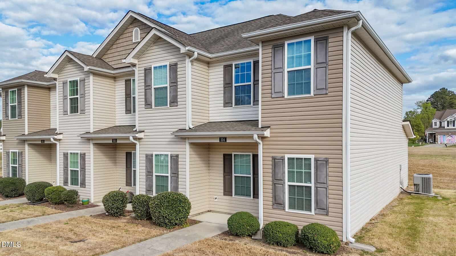 A two-story beige townhouse with white trim and gray shutters, featuring multiple windows, small front bushes, and a concrete walkway, set against a partly cloudy sky.