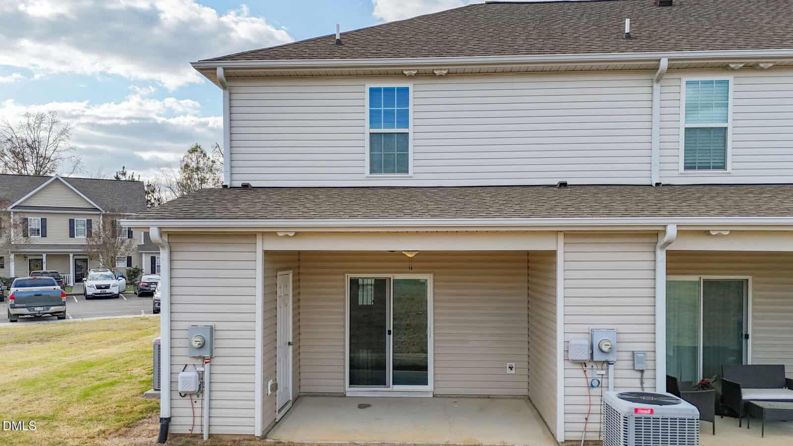 A two-story beige townhouse with a small concrete patio, sliding glass door, and an upper window. HVAC units and utility meters are visible near the patio. Neighboring homes and parked cars are in the background.
