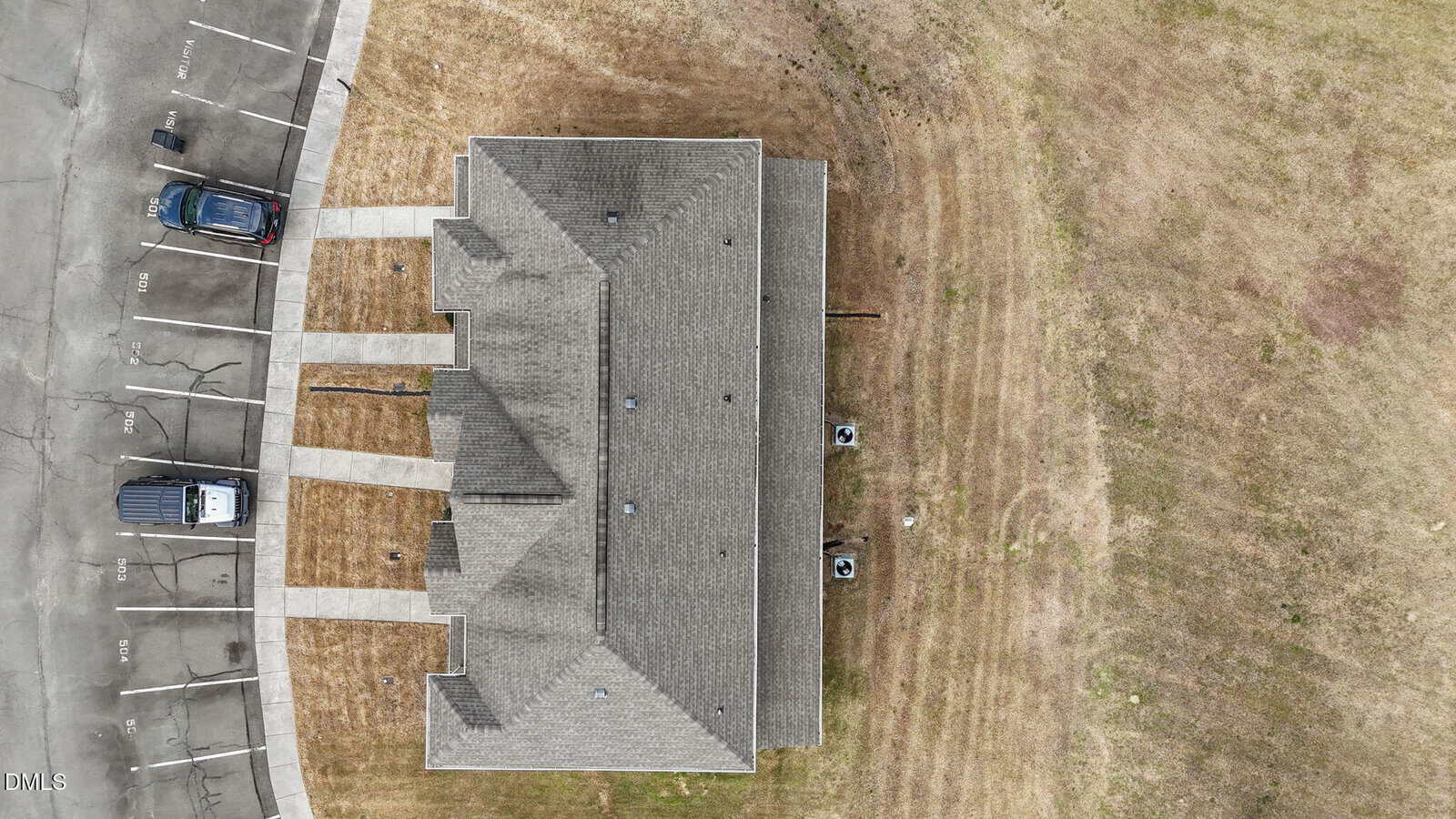 Aerial view of a rectangular building with a gray roof, two parked vehicles on the left, and surrounding brown and green grass. Sidewalks lead to the entrances on either end of the building.