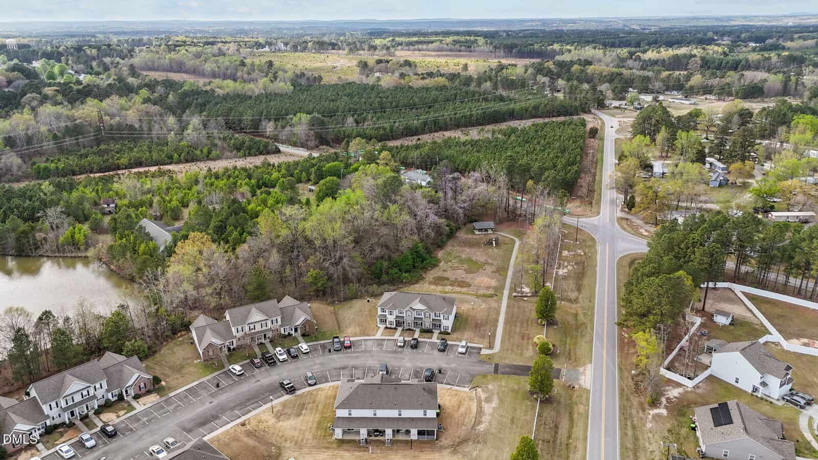 Aerial view of a suburban neighborhood with houses, a parking lot, a pond, and dense green forests in the background under a partly cloudy sky.