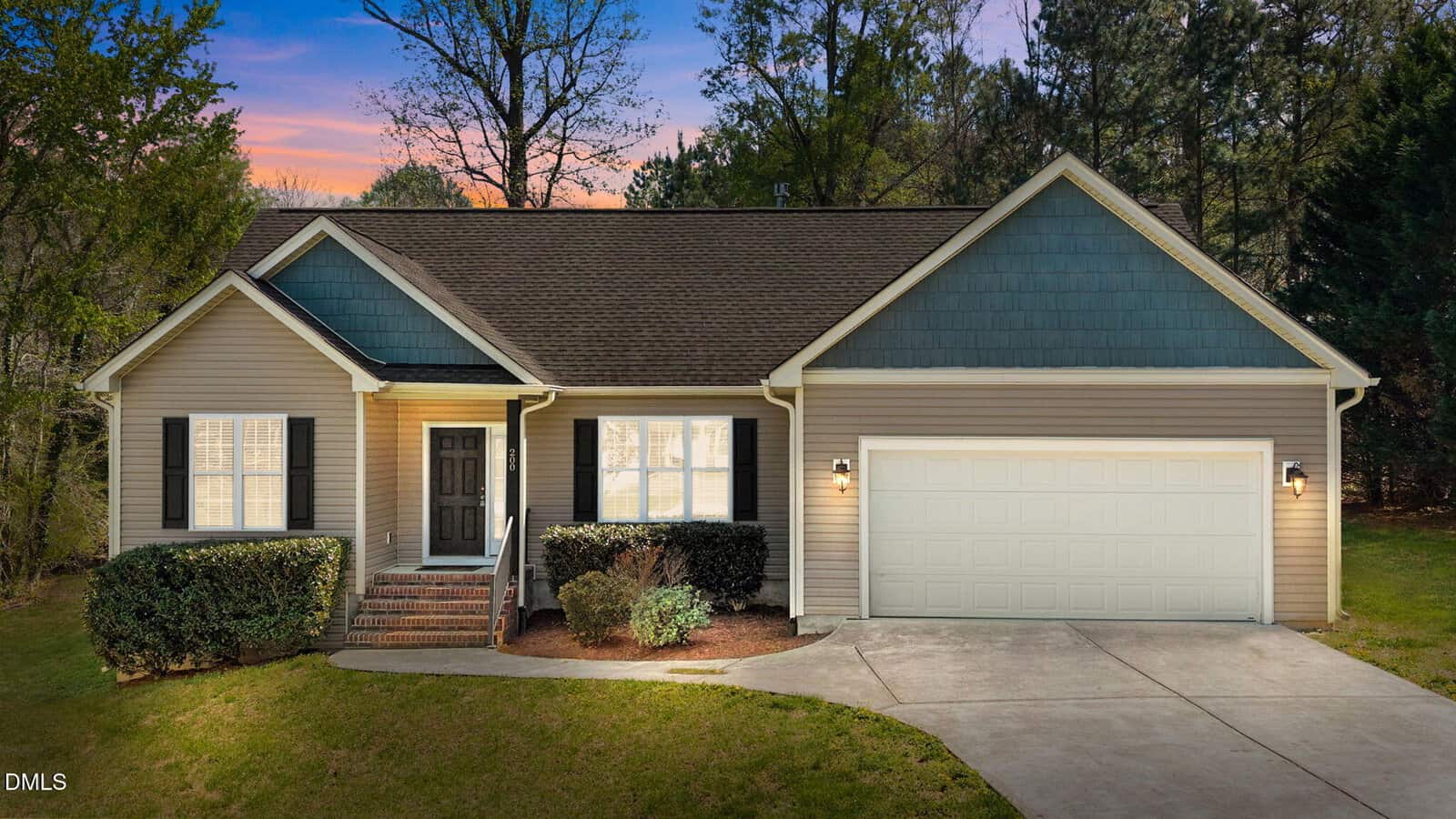 A single-story house with beige siding, dark shutters, a front porch with steps, a double garage, and a driveway. The yard has trimmed bushes and trees in the background, and the sun is setting in the sky.