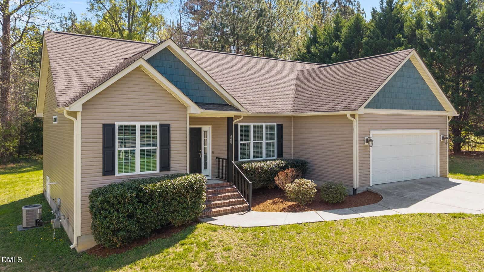 Single-story beige house with white trim, dark shutters, a front porch with brick steps, two-car garage, manicured shrubs, and a concrete driveway set against a backdrop of trees and grass.