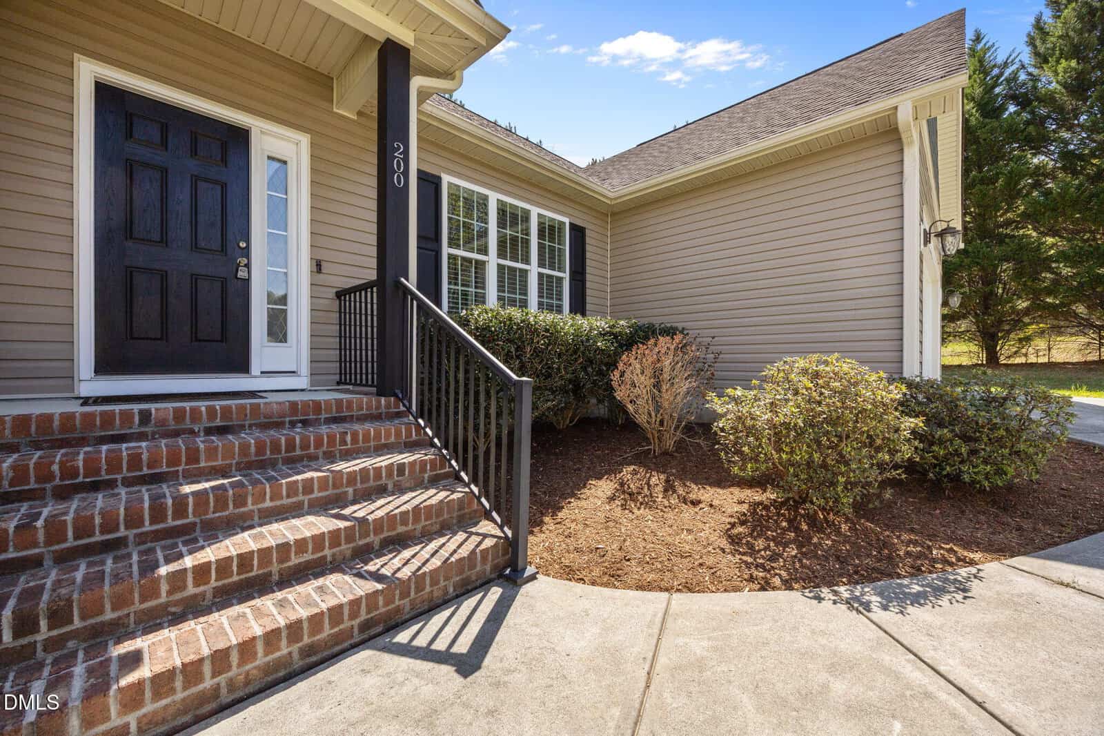 A brick staircase with black railings leads to a black front door of a beige house with white trim. Well-kept bushes line the mulched landscaping next to a concrete walkway.
