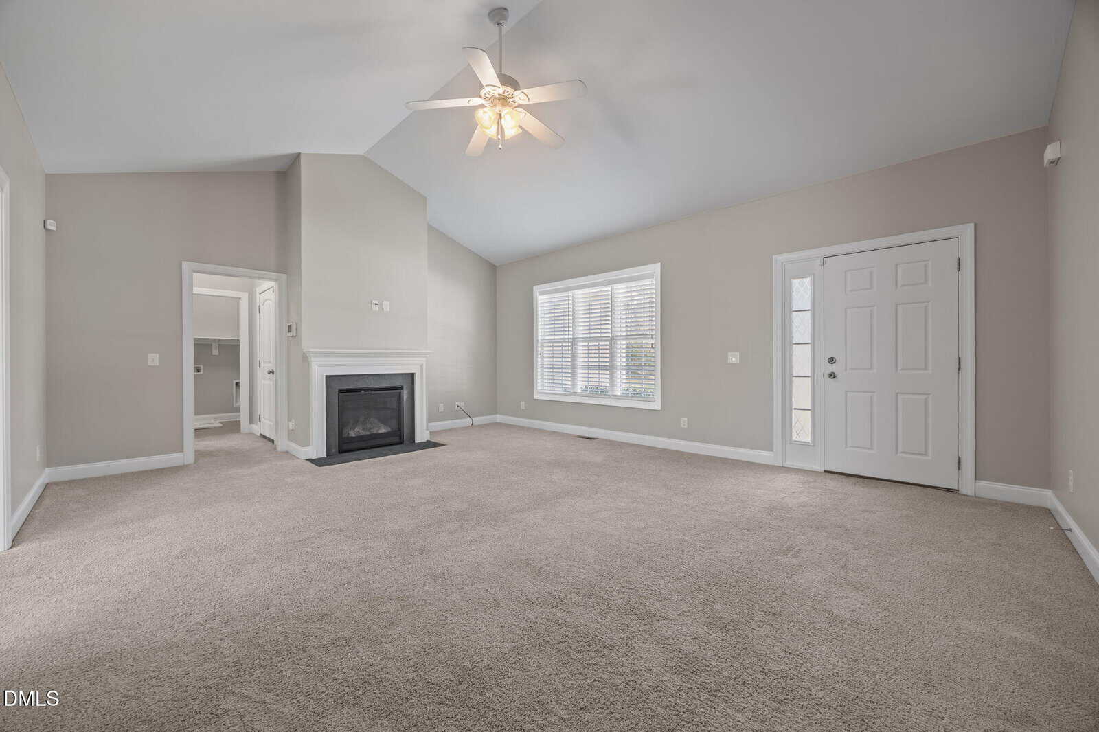A spacious, empty living room with beige carpet, light gray walls, a ceiling fan, a fireplace with a mantel, a white front door, and a large window with white blinds letting in natural light.