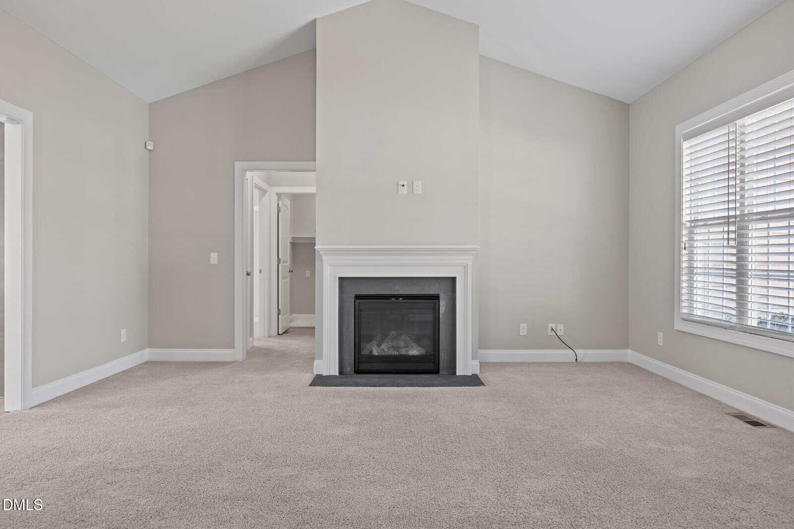A carpeted living room with beige walls, a white-trimmed fireplace centered on one wall, two open doorways on the left, and a large window with white blinds on the right. The room is empty and well-lit.